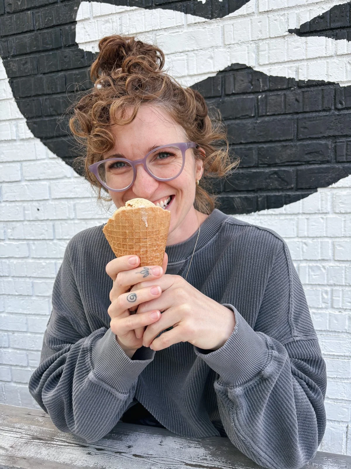 A woman with curly hair, glasses, and tattoos on her fingers smiling and holding an ice cream cone, sitting in front of a white brick wall with graphic black and white art.