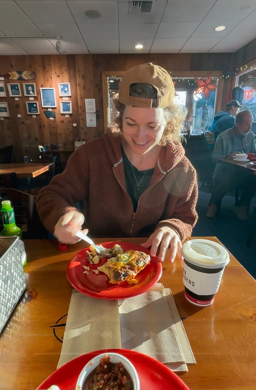 A woman in a tan cap and brown hoodie smiling as she eats pizza at a wooden table in a cozy restaurant. There is a cup of coffee and bottles on the table, with other diners in the background and holiday decorations outside the window.