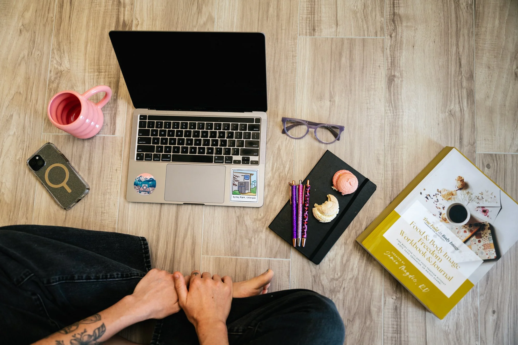 A top-down view of a workspace on a wooden floor, including a laptop, pink mug, smartphone, glasses, notebooks, pens, a book about food and body image, and a person's hands and legs in jeans.