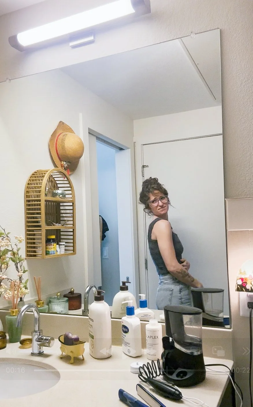 A woman with glasses and curly hair, wearing a gray tank top and jeans, stands in front of a bathroom mirror. The bathroom counter has bottles of lotion, soap, and other personal care items, along with a small plush toy and a blender. A hat hangs on the wall, and there are decorative plants and small items on the counter.