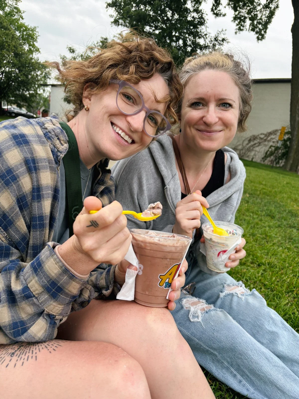 Two women sitting outside on grass, smiling, holding cups of ice cream, one with a container of chocolate ice cream and the other with vanilla and rainbow sprinkles.