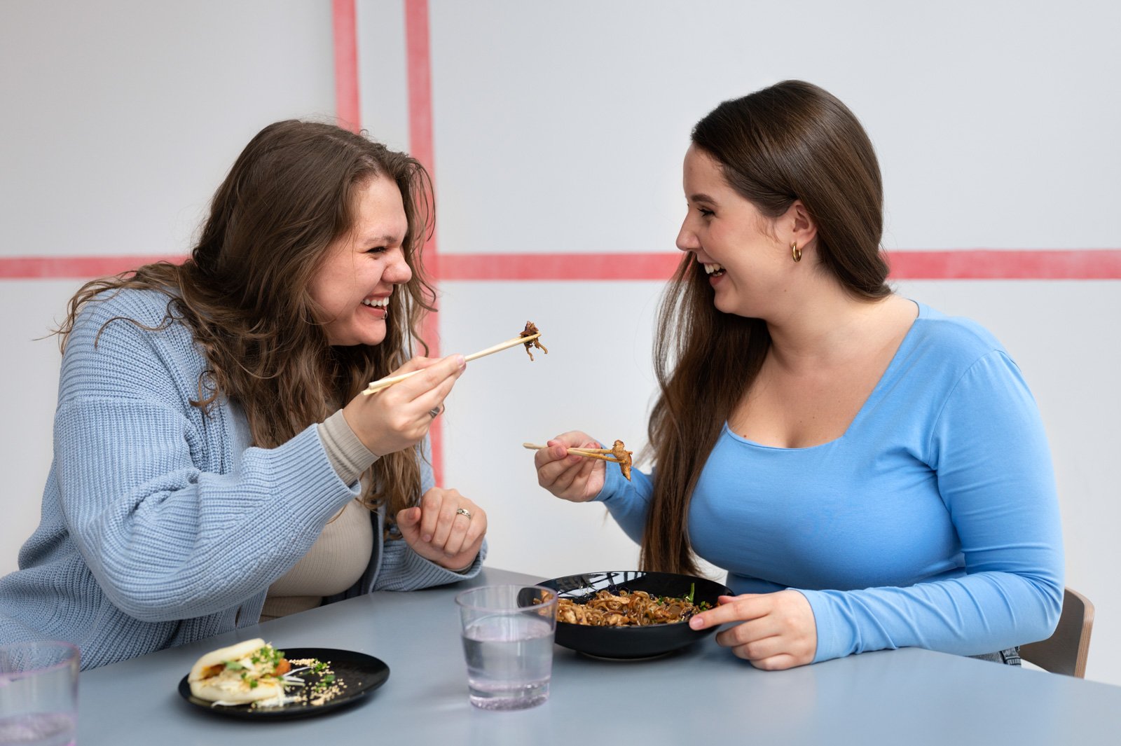 Two women enjoying a meal together, sharing a laugh as one offers the other a piece of food with chopsticks.