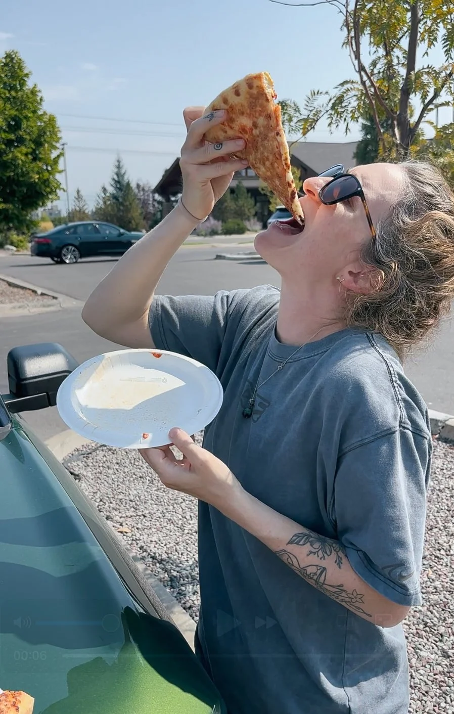 A woman with sunglasses is outside, holding a slice of pizza near her mouth and about to take a bite. She is holding a paper plate with some food remnants. She has tattoos on her left arm and is wearing a grey T-shirt.