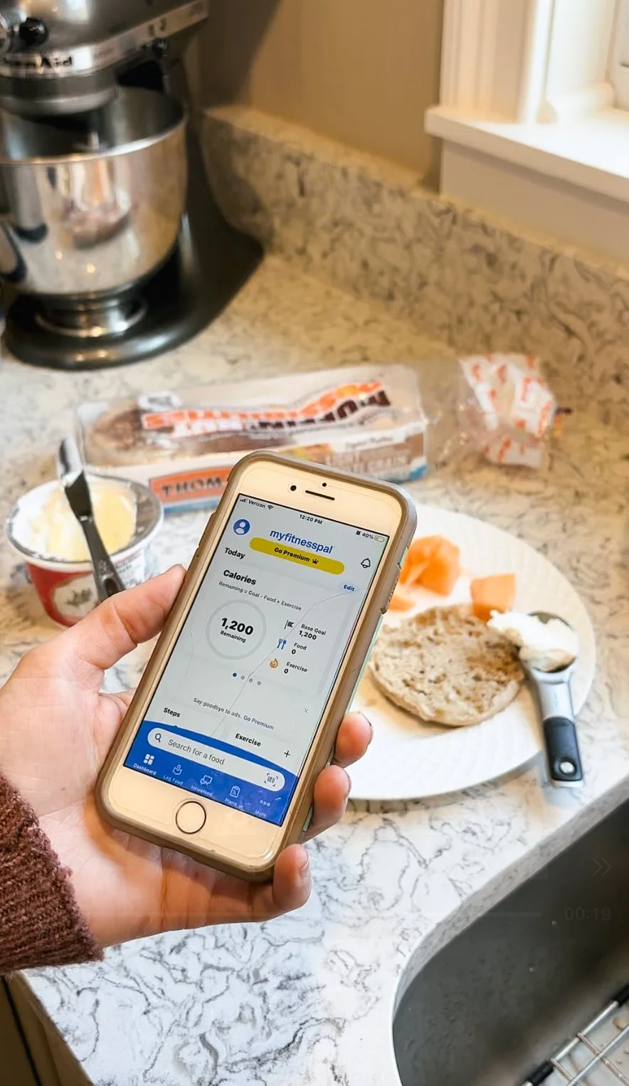 A kitchen countertop with a sandwich on a paper plate, a yogurt container with a spoon, a loaf of bread, and a hand holding a smartphone showing a fitness app. There is also a stand mixer and some packaged bread in the background.
