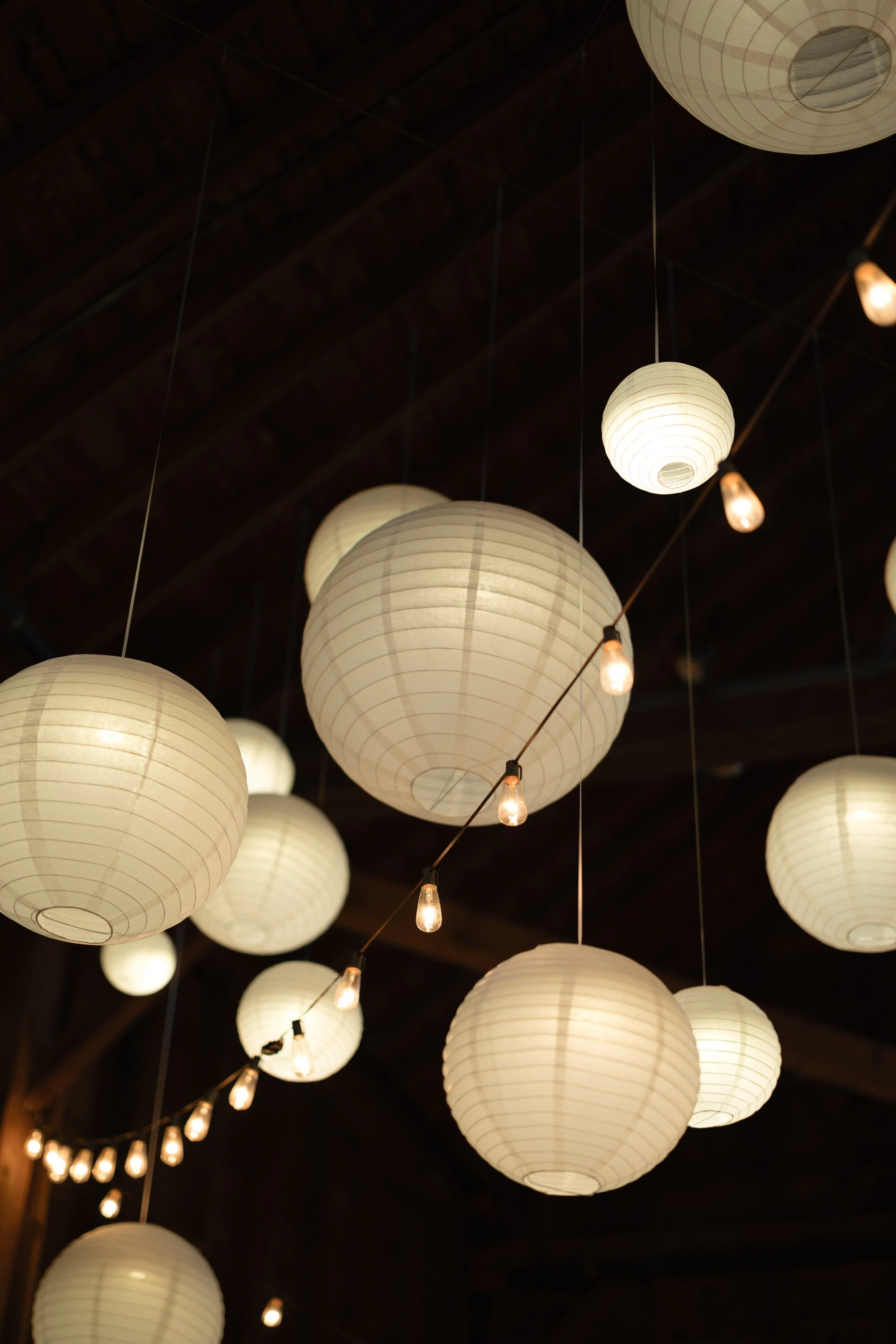 Close-up night view of glowing white paper lanterns installed in the barn rafters at Gedney Farm by Berkshire Event Lighting.