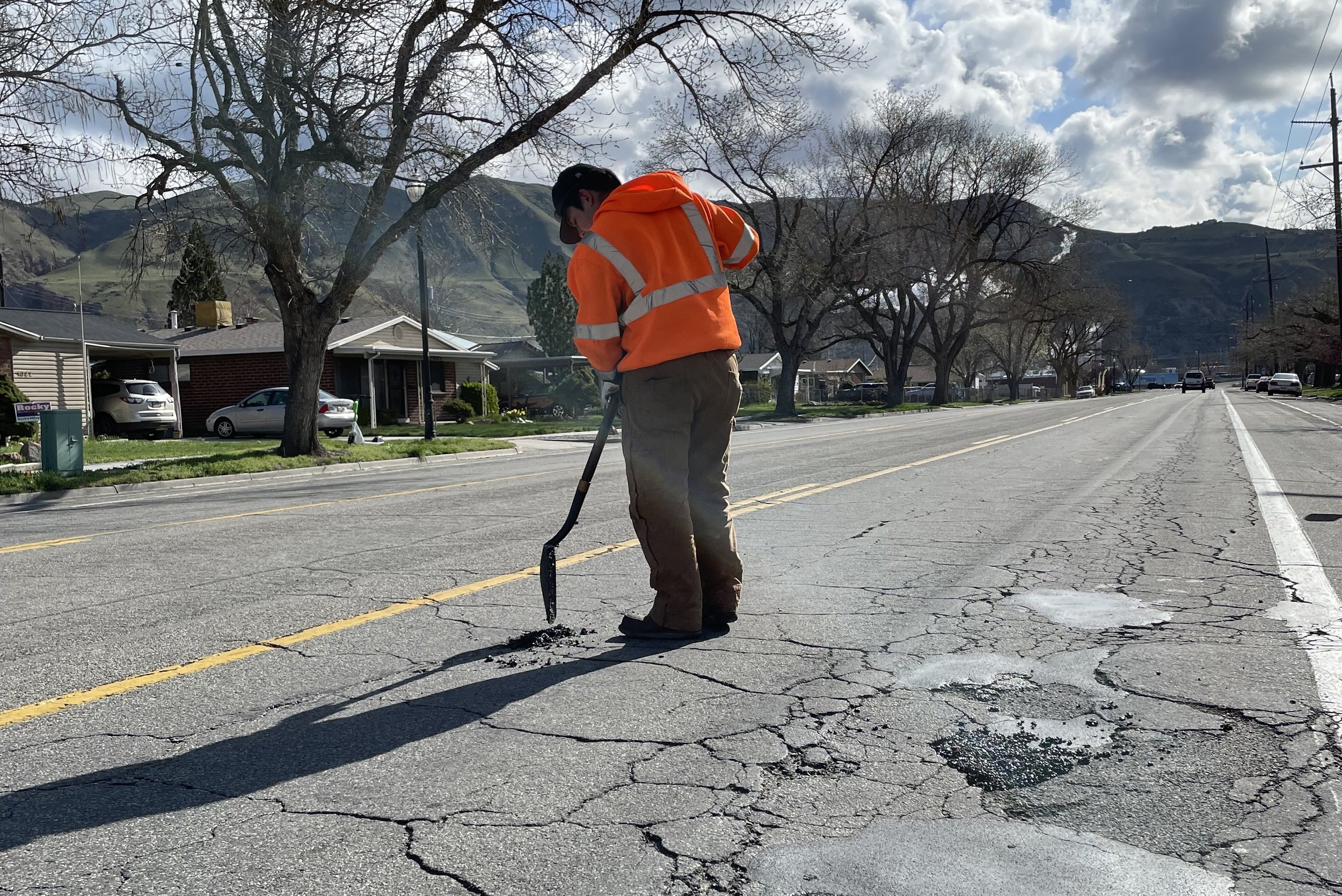 A city worker in an orange vest fills a pothole using a shovel and asphalt.