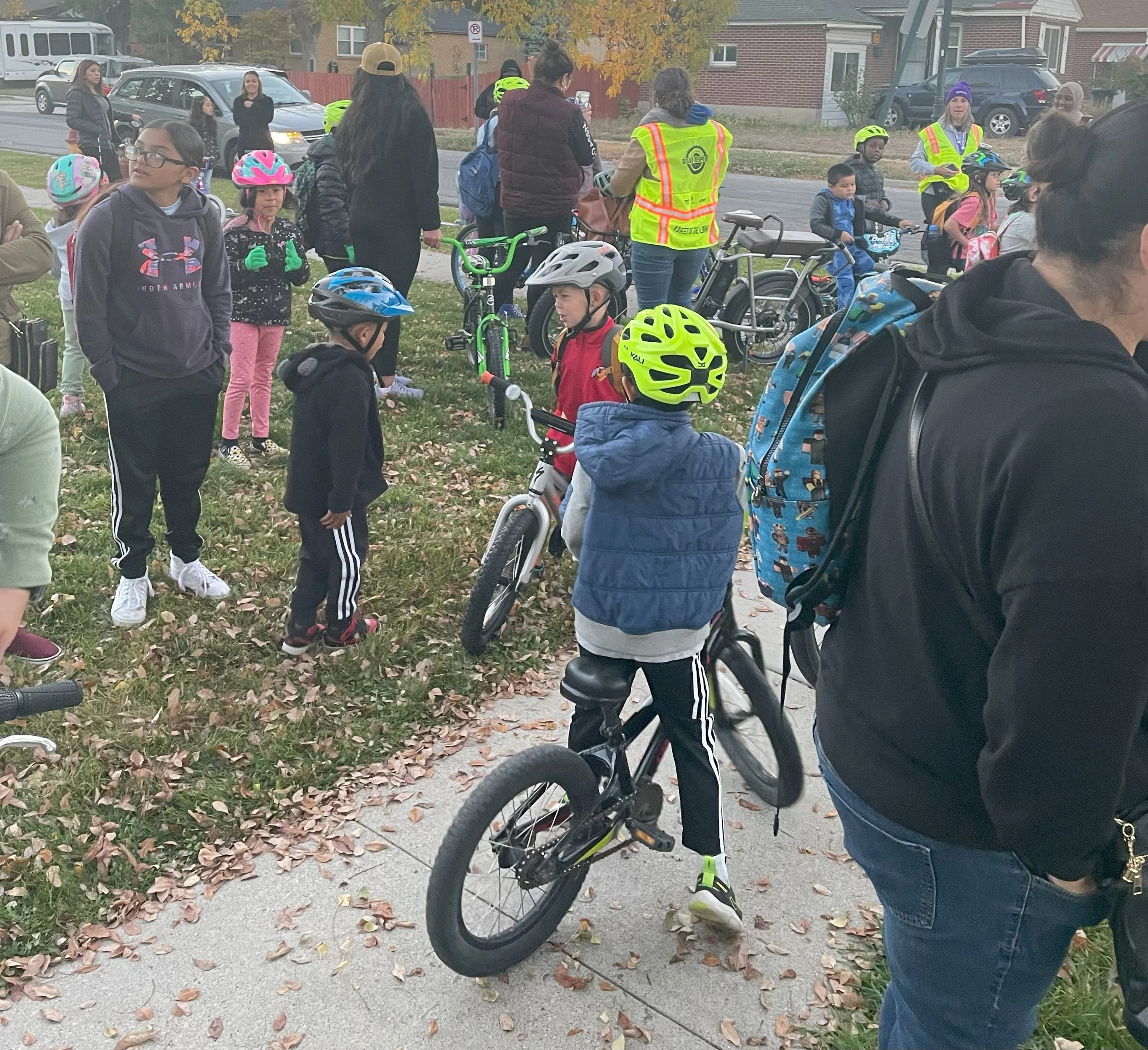Children and adults arrayed in cycling equipment on a leaf-strewn lawn, preparing to ride to school.