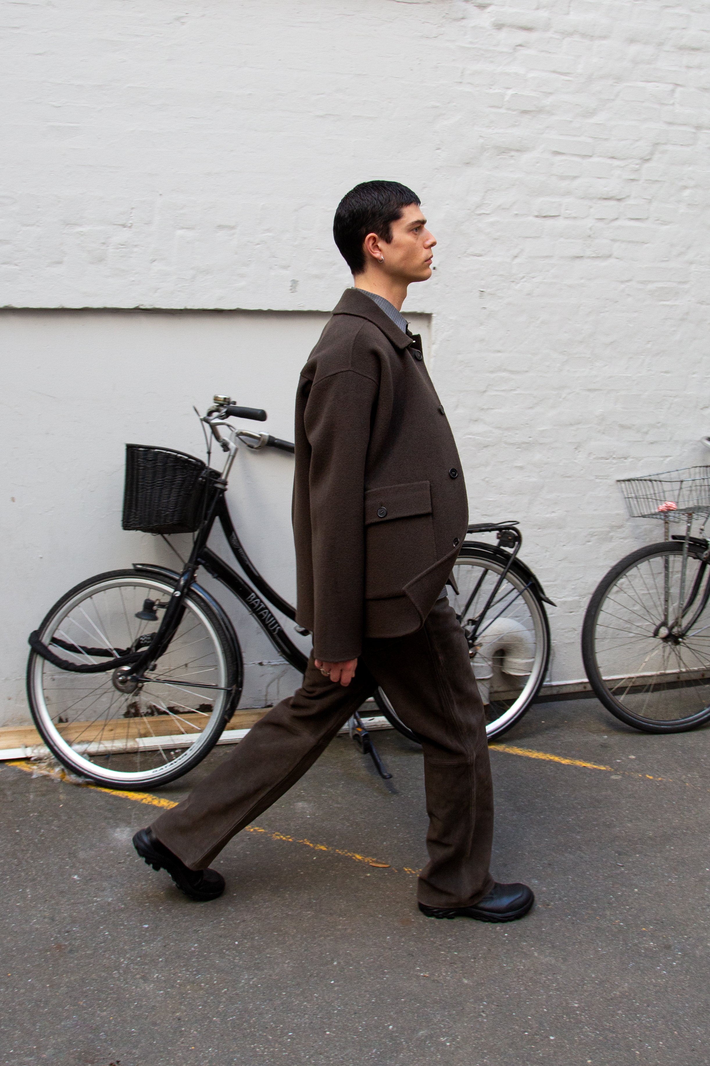 A young man with dark hair and light skin walking past a white brick wall, wearing a dark brown blazer, brown pants, and black shoes, walking on an asphalt driveway with bicycles in the background.