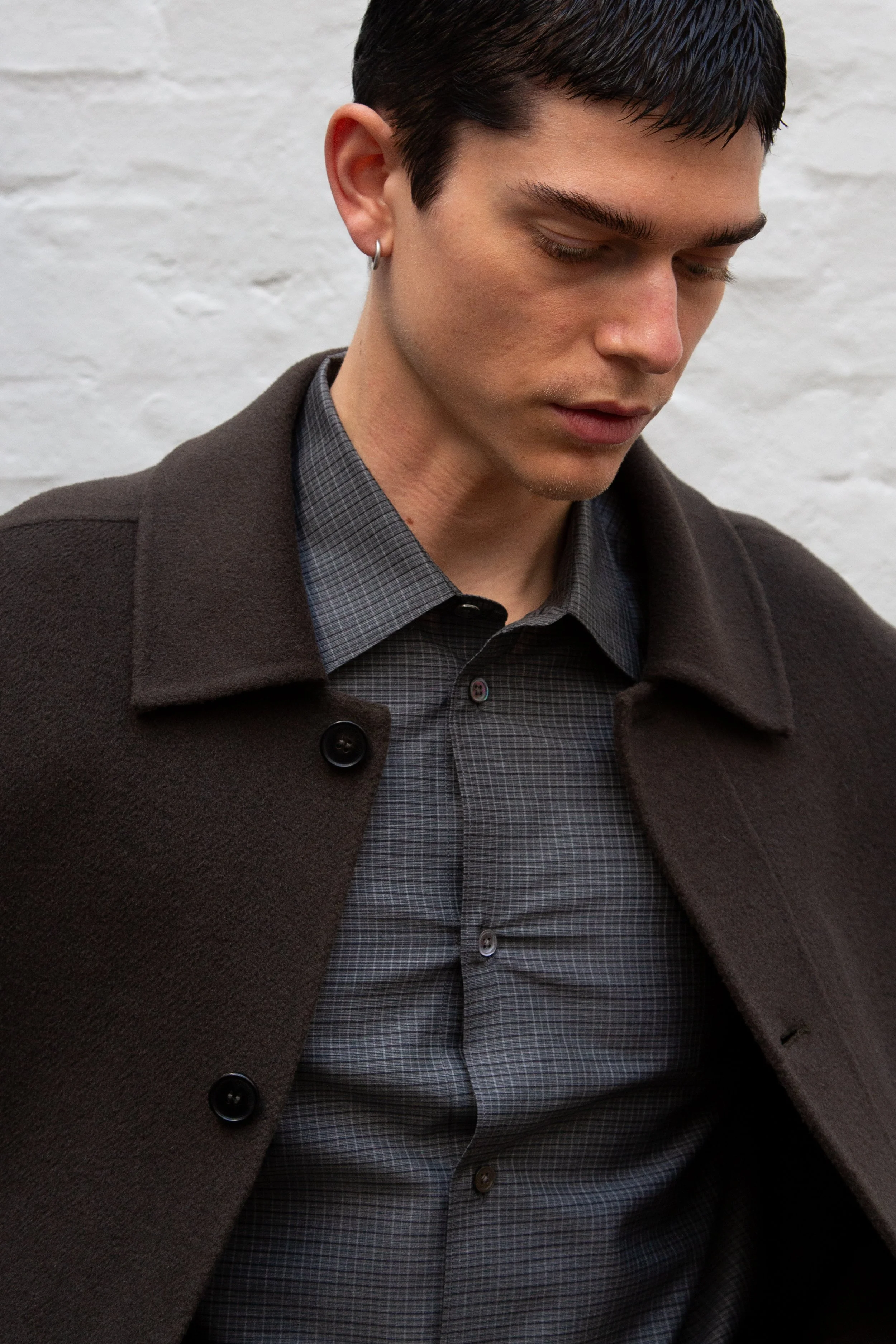 A young man with short black hair and an earring, wearing a dark jacket over a gray checkered shirt, looking downward against a white brick wall background.