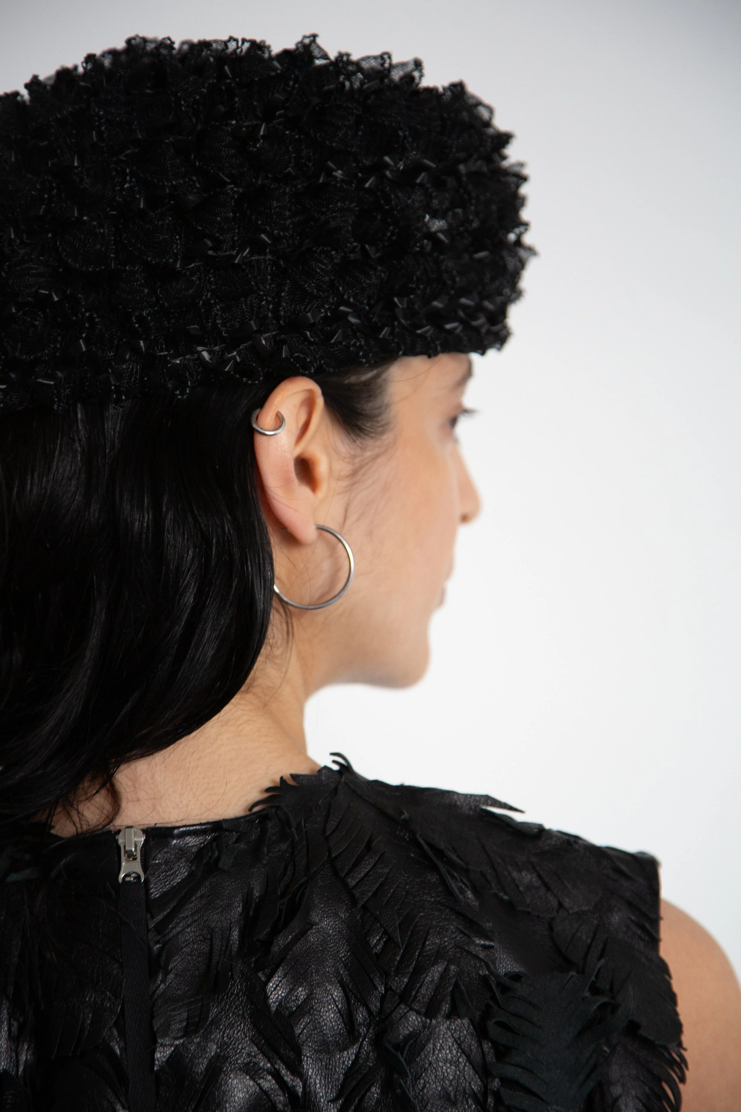 A woman viewed from the side, wearing a textured black headpiece and earrings, with a black feathered top, against a plain white background.