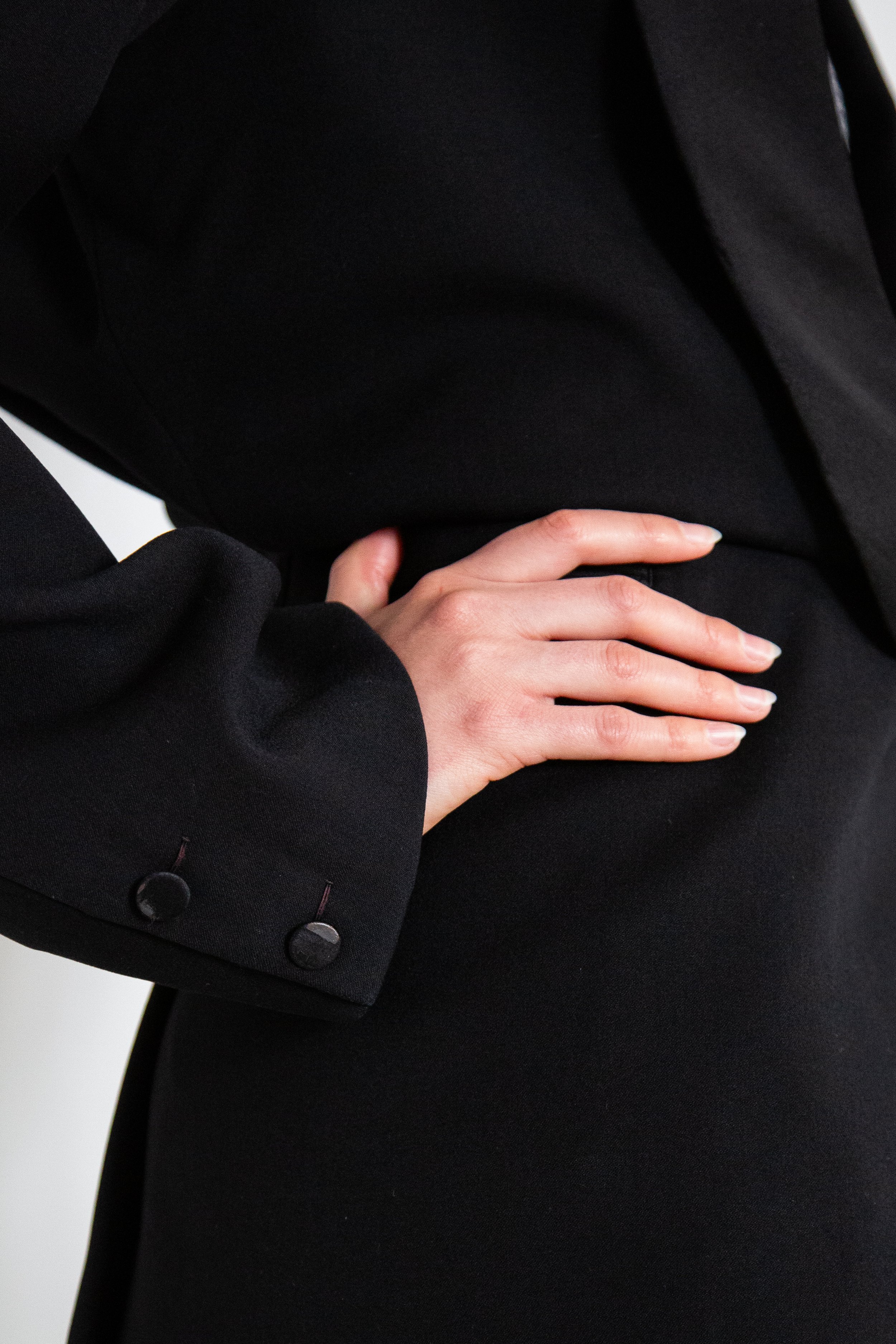 A person's hand with manicured nails resting on their hip, dressed in a black blazer.