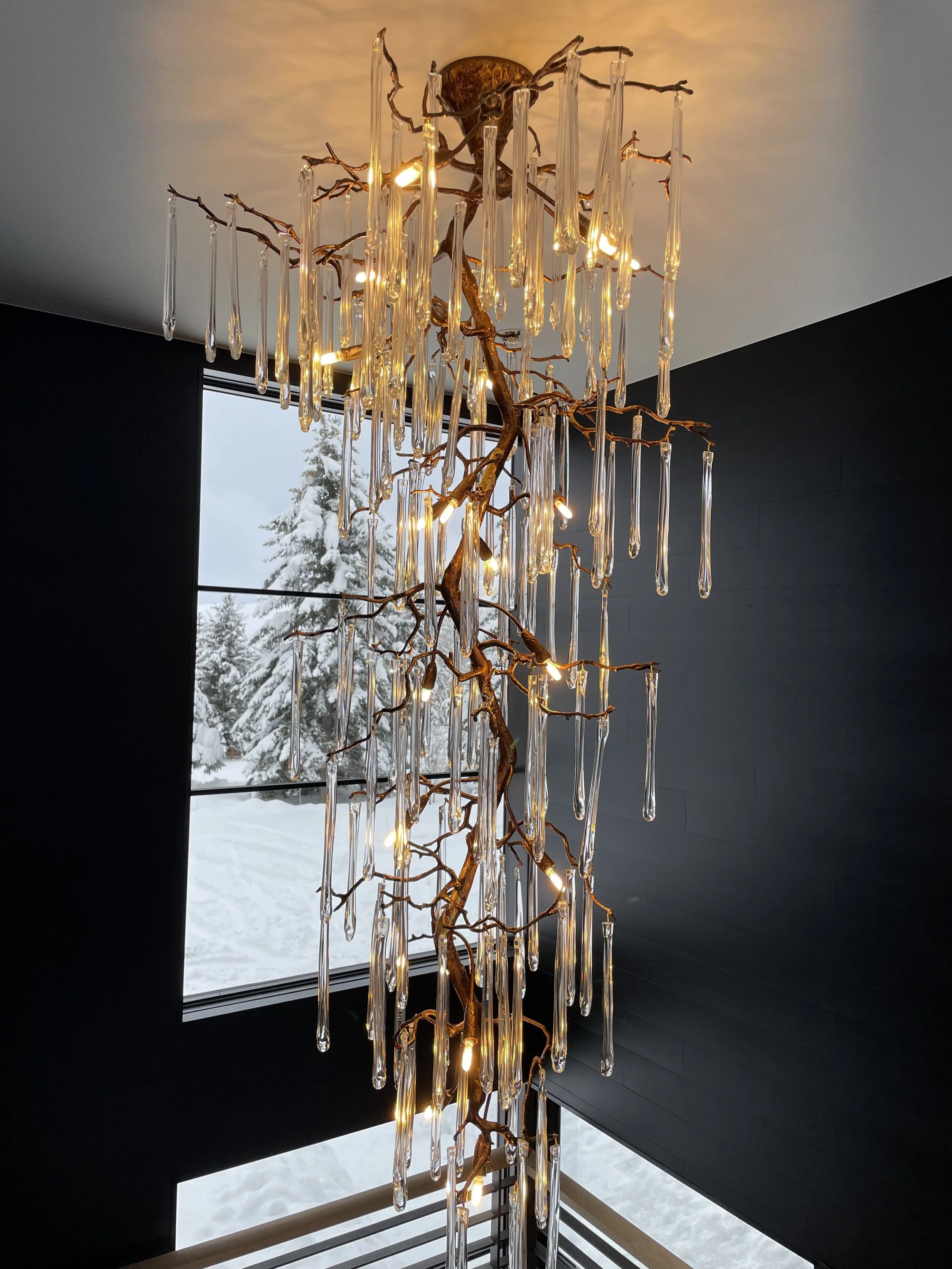 Elegant chandelier with a branch-like structure and hanging glass icicle-shaped ornaments, illuminated inside a modern home with dark walls and snowy landscape outside