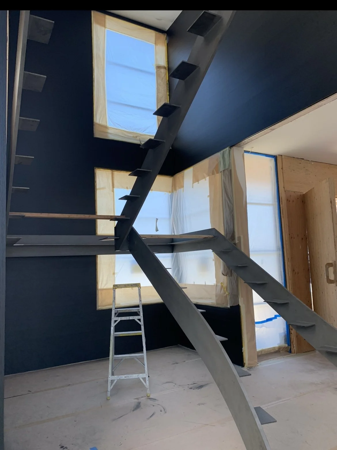 Interior of a house under construction with stairs, windows covered with protective plastic, and a ladder. The walls and ceiling are partially finished.