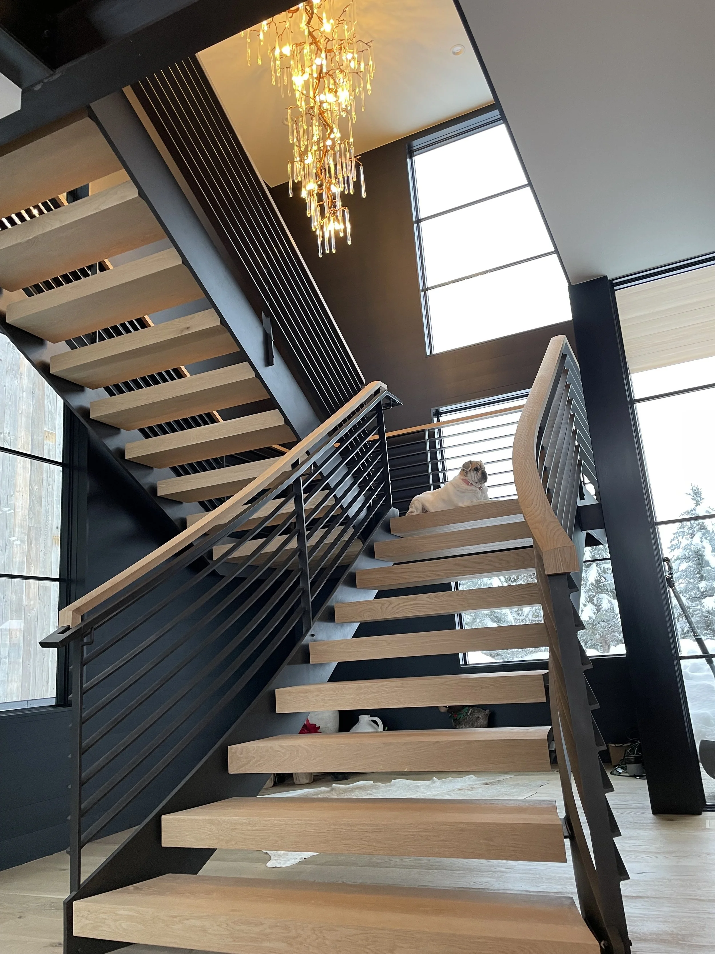 Interior view of a modern staircase with wooden steps and black metal railings, leading up to a loft area with large windows, a cat resting on the top step, and a chandelier hanging from the ceiling.
