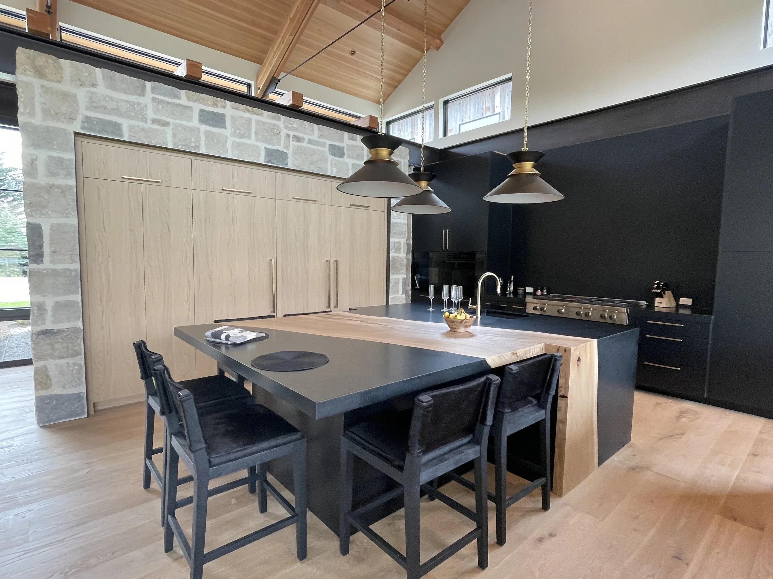 Modern kitchen with black and wooden elements, featuring black dining chairs, a black kitchen counter with a waterfall edge, black ceiling, black cabinets, a large wooden kitchen island, hanging pendant lights, and a stone wall with large windows.