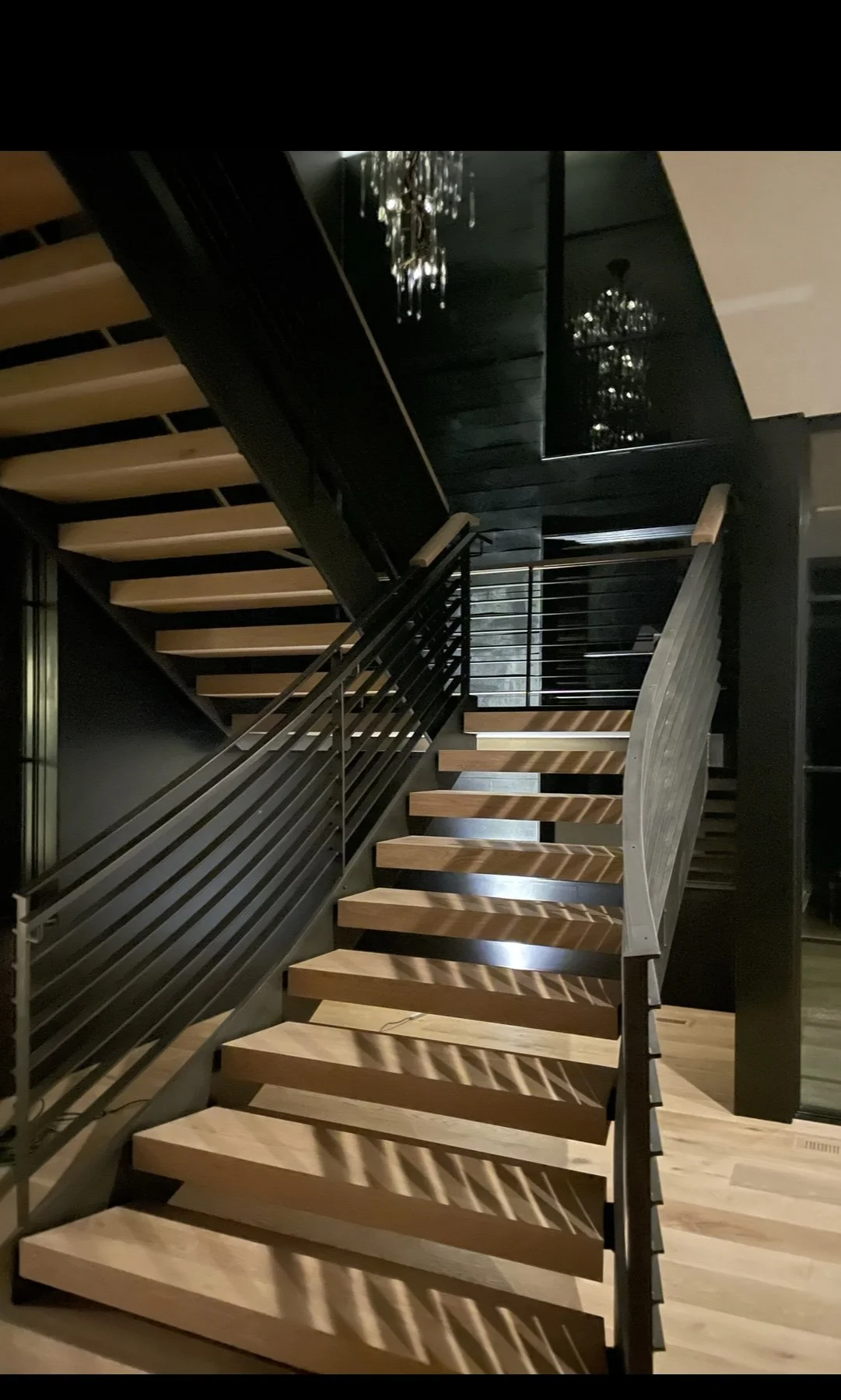 Modern staircase with wooden steps, metal railing, and black walls, reflecting chandeliers in a sleek, contemporary interior.