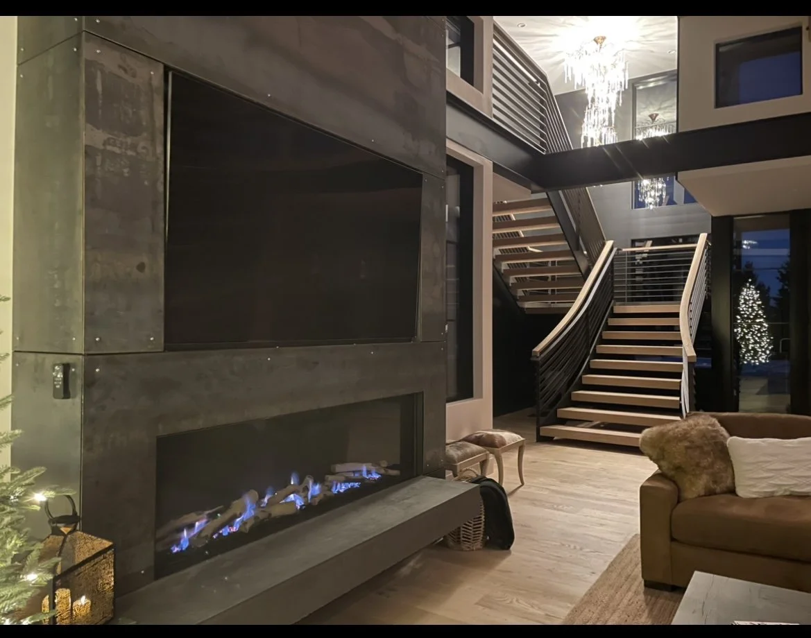 Modern living room with a flat-screen TV above a linear gas fireplace, a brown sofa with a fur throw, a staircase with a black railing, and a chandelier, decorated for Christmas with a lit Christmas tree outside the window.