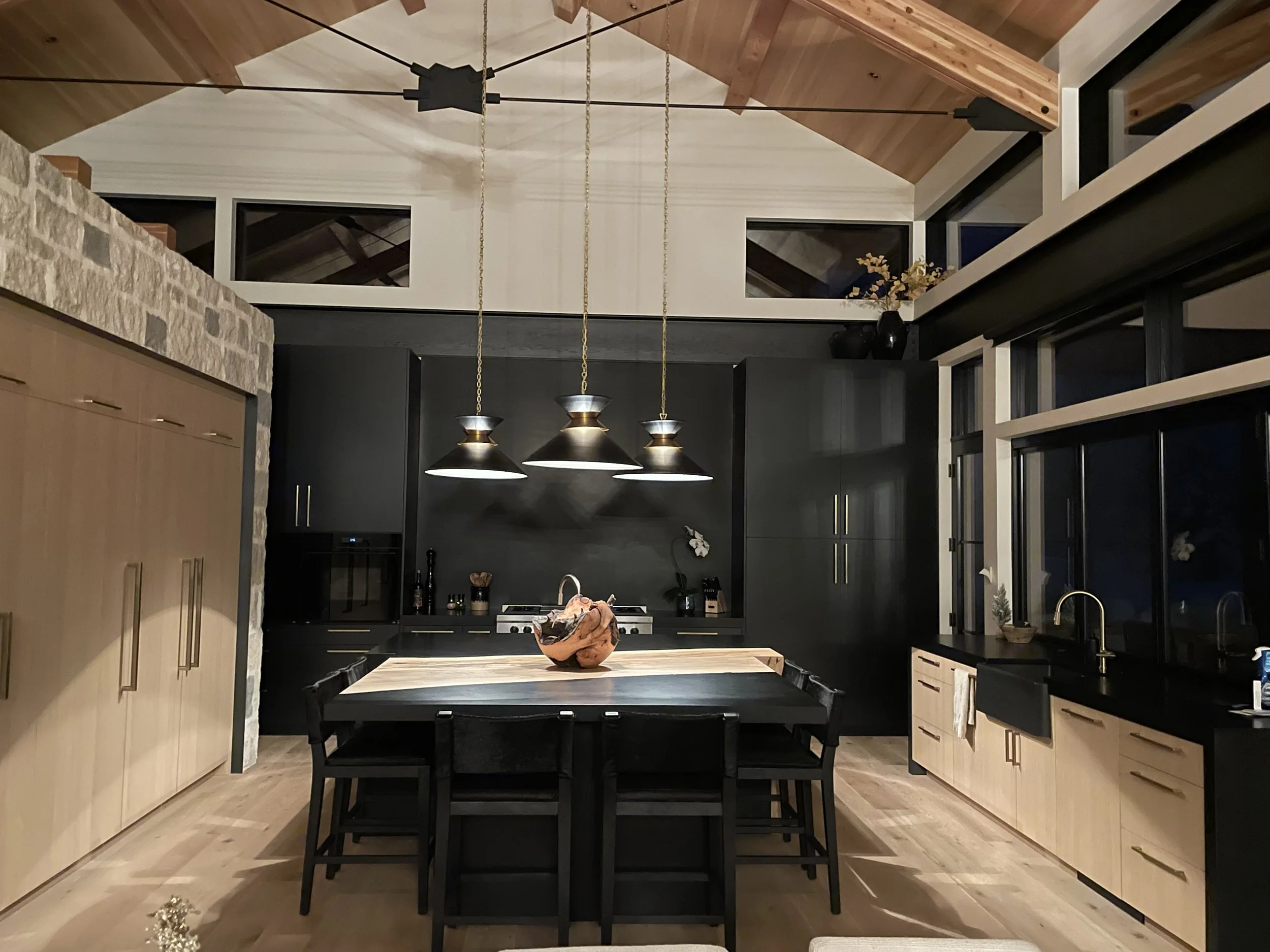 Modern kitchen with black and wooden cabinetry, stone accent wall, large black dining table with chairs, black pendant lights, and large windows at night.