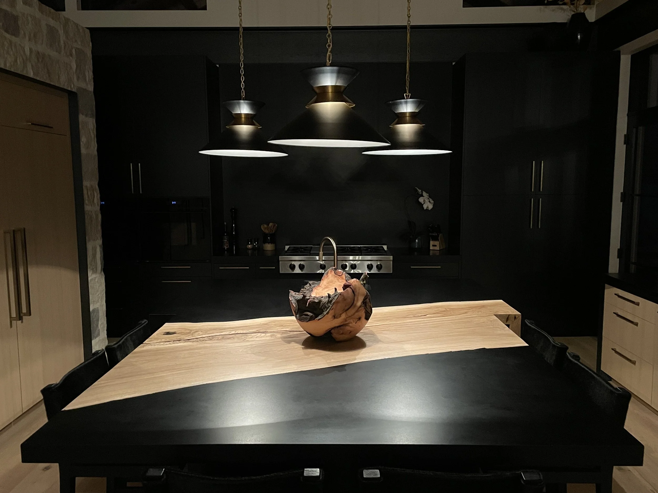A modern kitchen with dark cabinets, a wooden dining table, and a decorative bowl in the center of the table. Pendant lights hang above the table, and a stove is visible in the background.