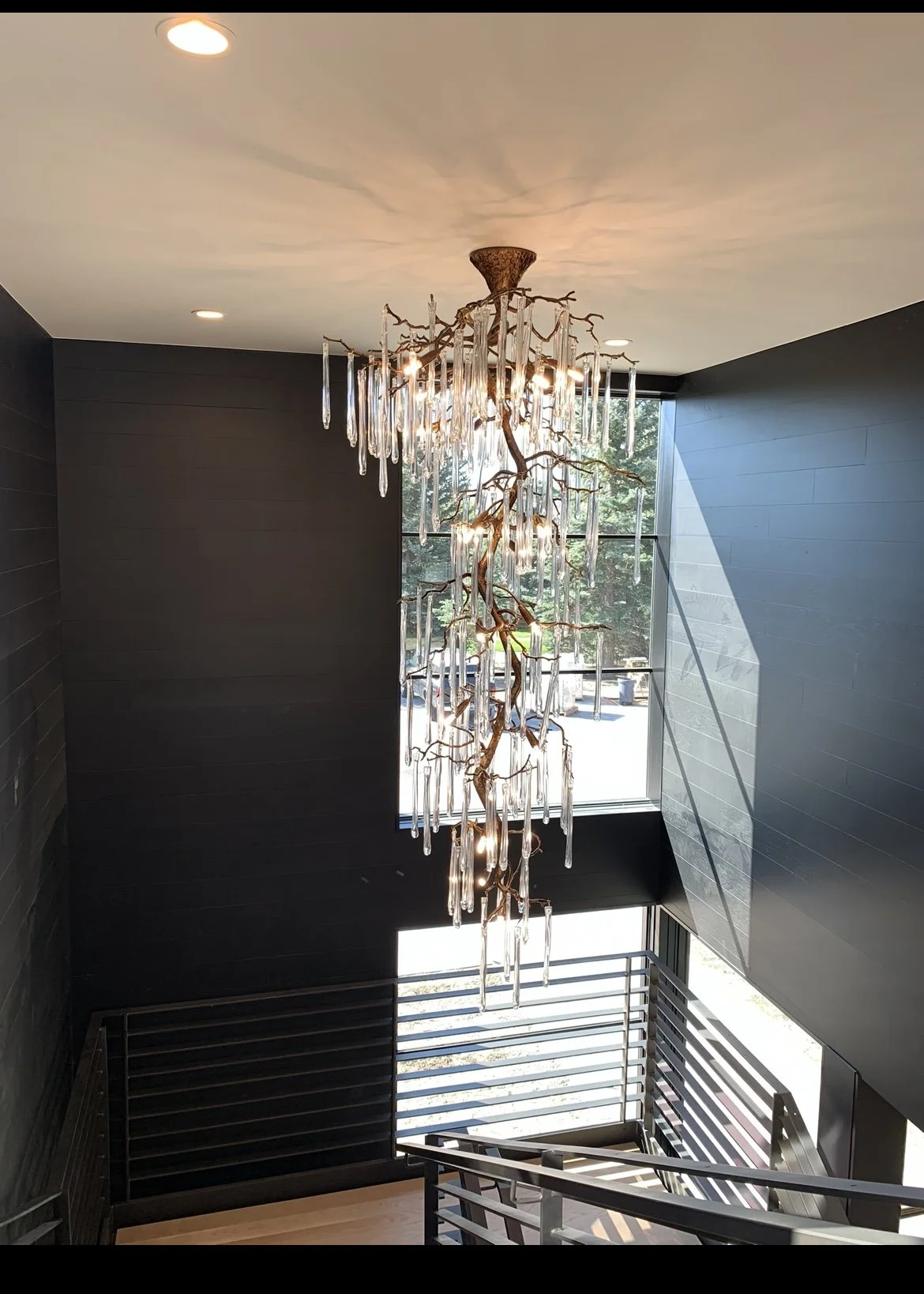 Modern stairwell with black walls, wooden railing, large window, and a decorative chandelier made of glass and metal resembling branches.