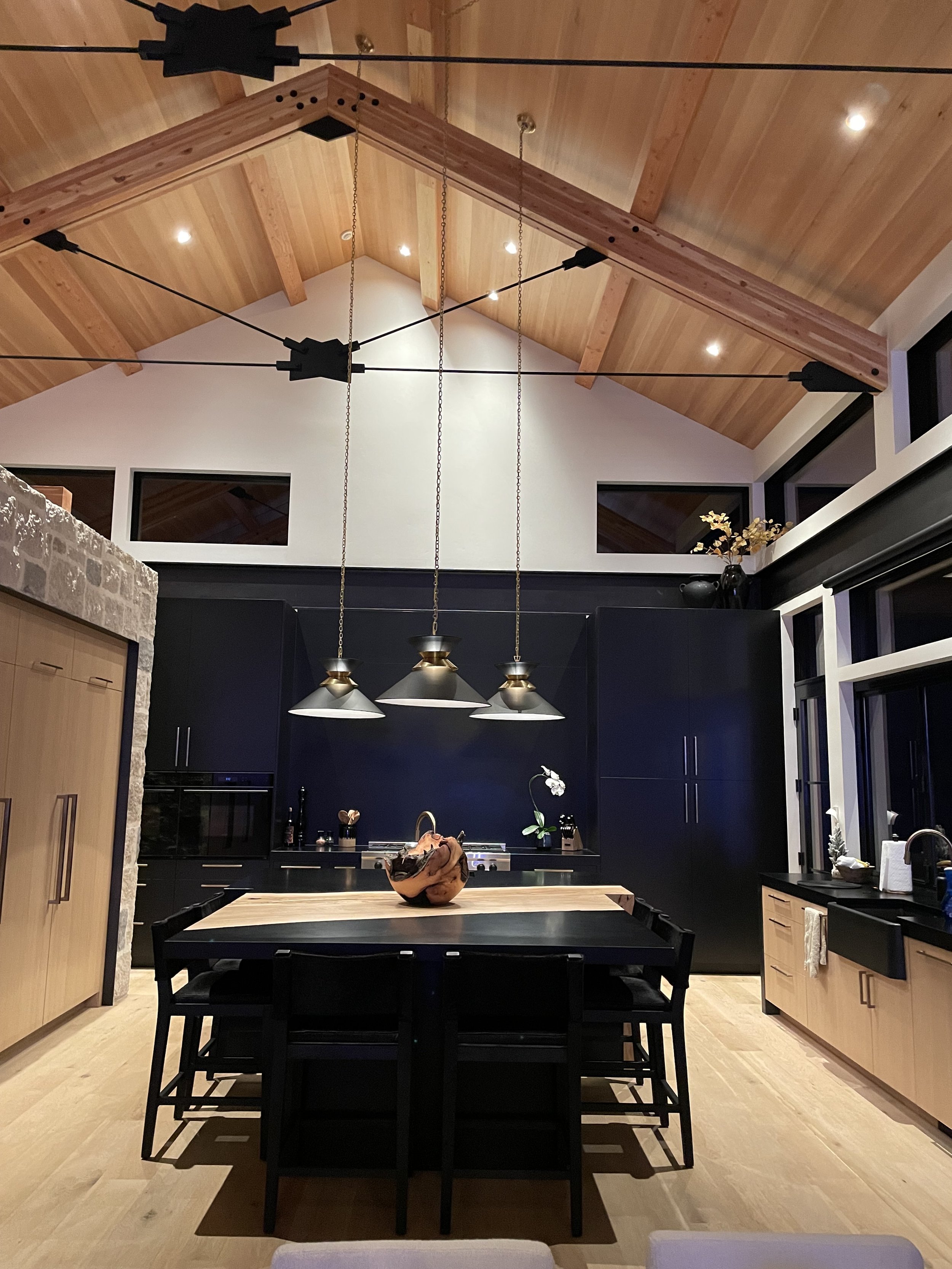 Modern kitchen with black cabinets, wooden island, and hanging pendant lights, featuring a high vaulted ceiling with exposed wooden beams and windows.