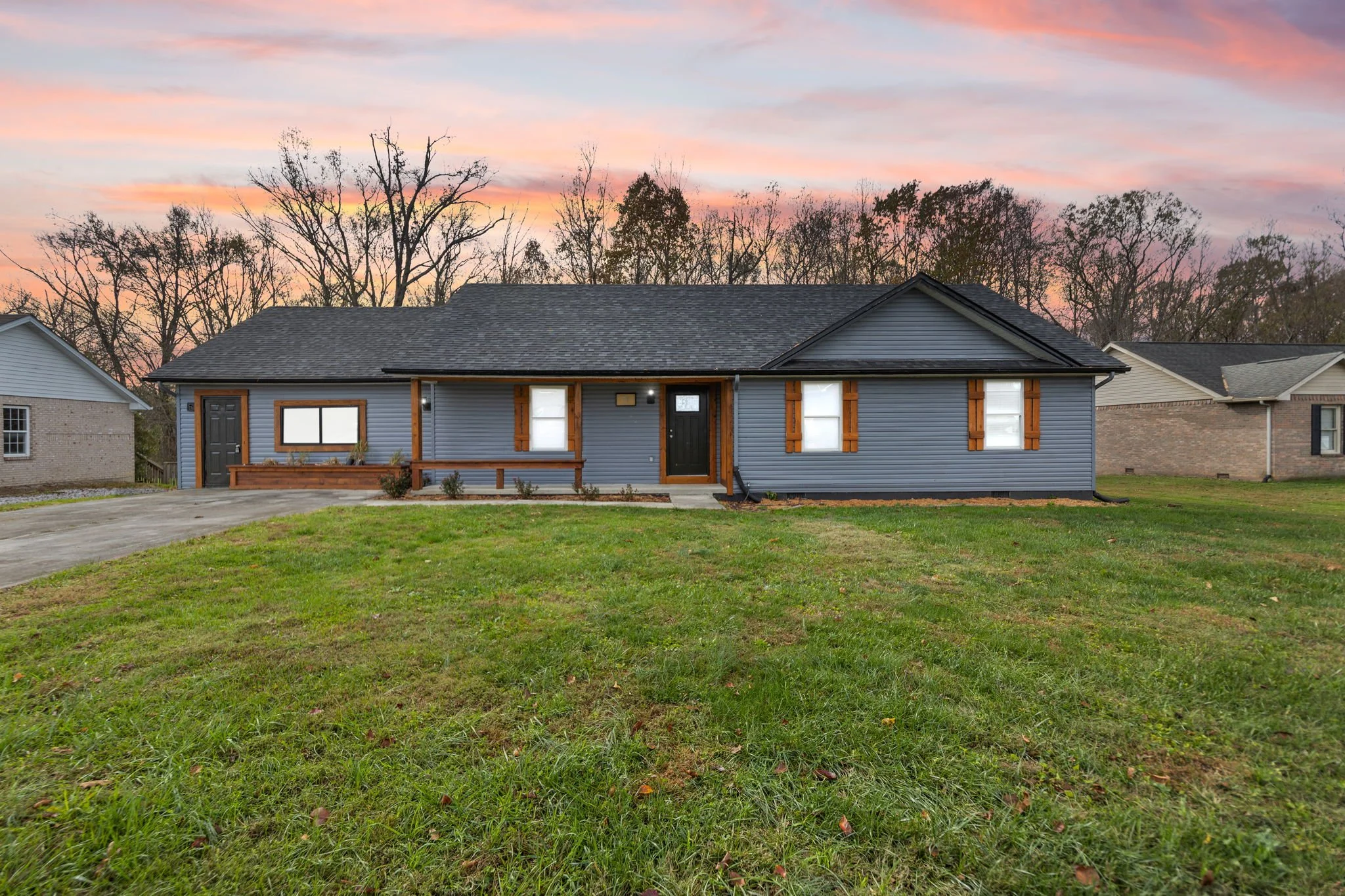 A single-story house with blue siding, wooden shutters, a small front porch, and a driveway, against a sunset sky and leafless trees in the background.