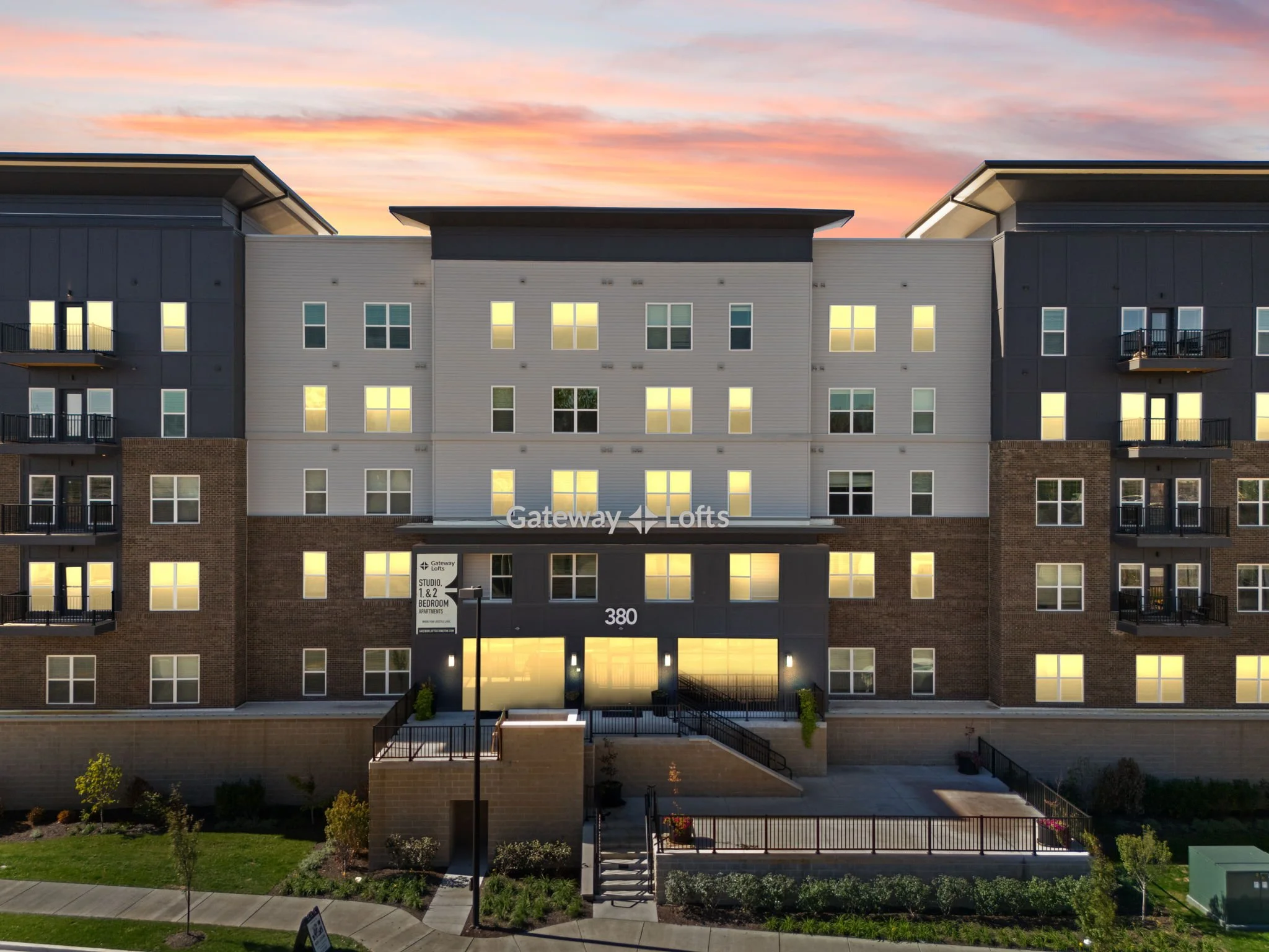 A modern multi-story apartment building during sunset with lit windows, balconies, and a landscaped front entrance. The sign reads 'Gateway Lofts'.