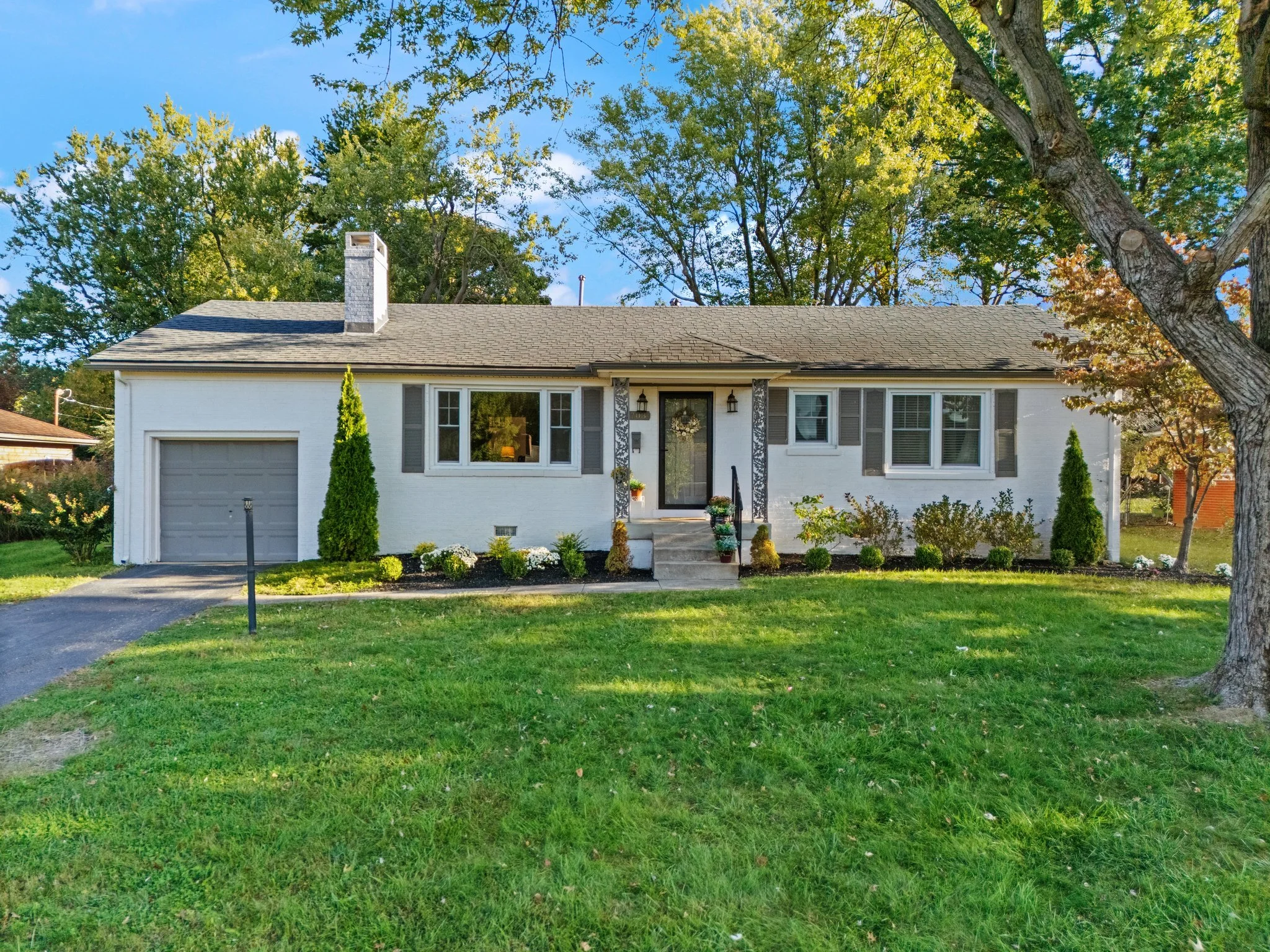 White house with gray shutters, a single garage door, chimney, and front steps, surrounded by green lawn and trees in the background.