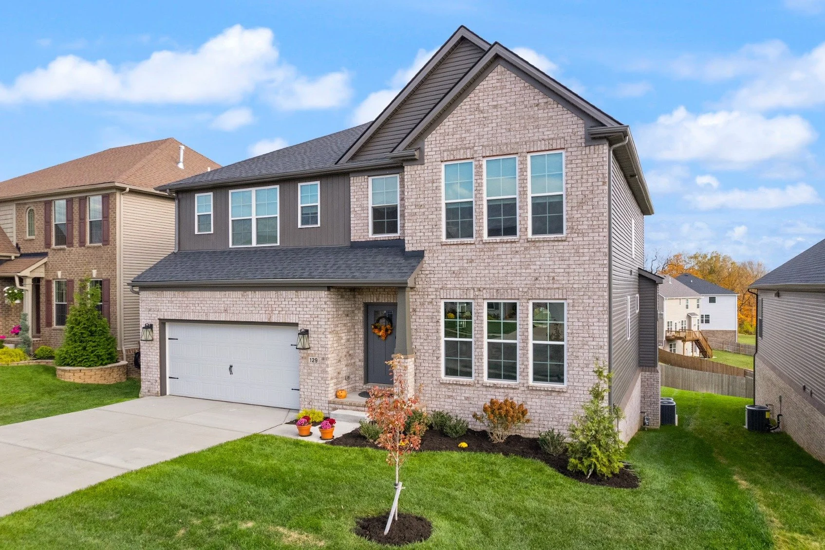 Modern two-story house with a brick and gray siding exterior, attached garage, and a well-maintained lawn with small trees and potted plants in fall decorations.