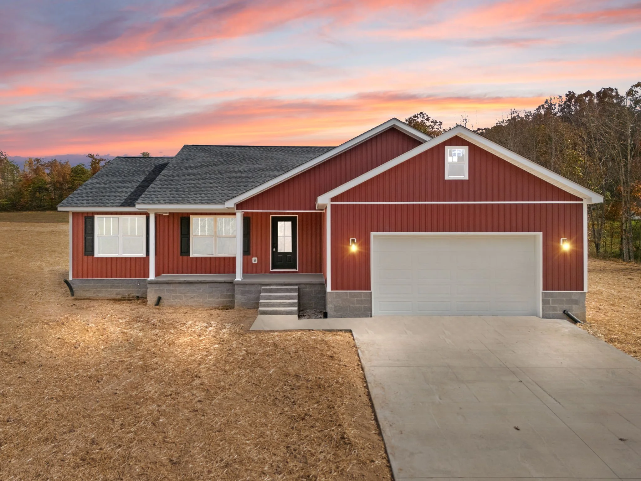 A newly constructed red house with white trim and a two-car garage, set against a sunset sky and a wooded background, with a concrete driveway leading to the garage.