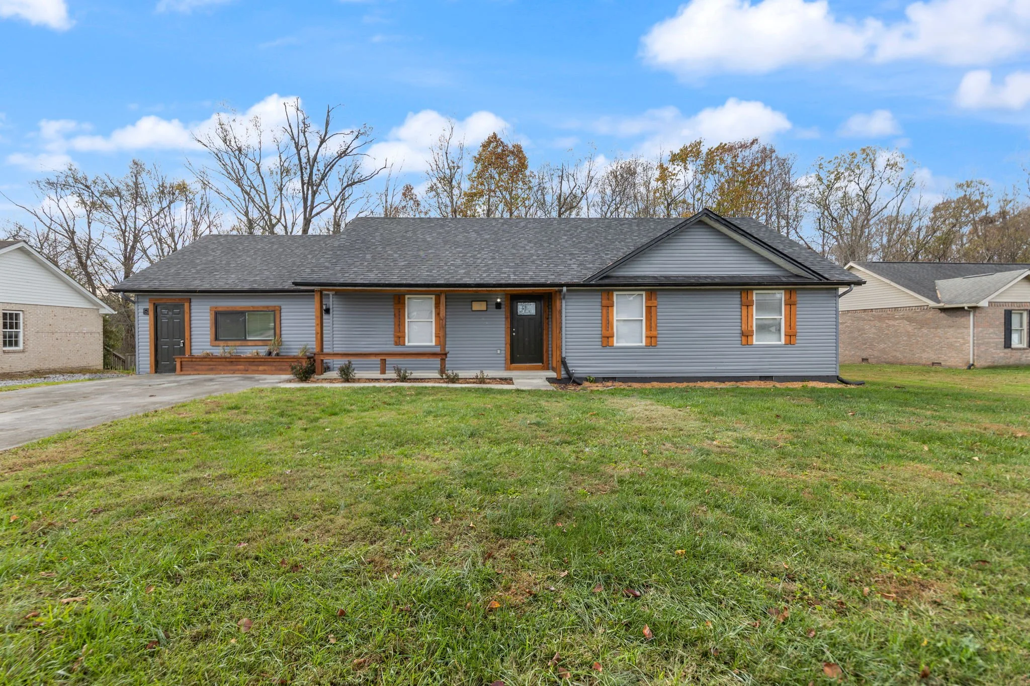 Single-story house with blue siding, brown window shutters, a black front door, and a small front porch, surrounded by a green lawn and other houses in the background under a partly cloudy sky.
