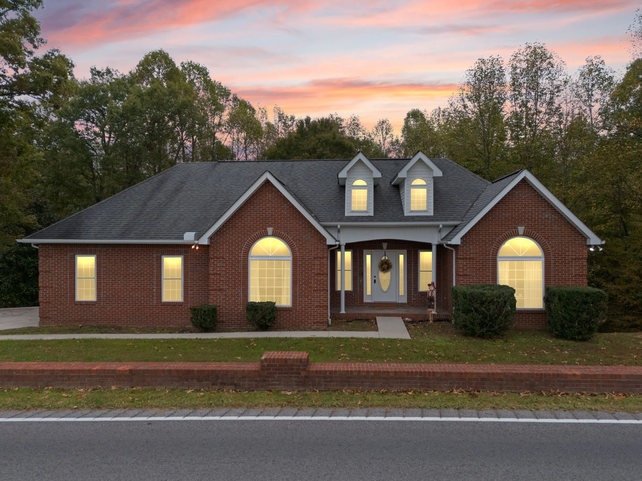 A brick house with five illuminated windows, an arched front door with a wreath, and a small porch with a snowman decoration, set against a sunset sky and surrounded by trees.