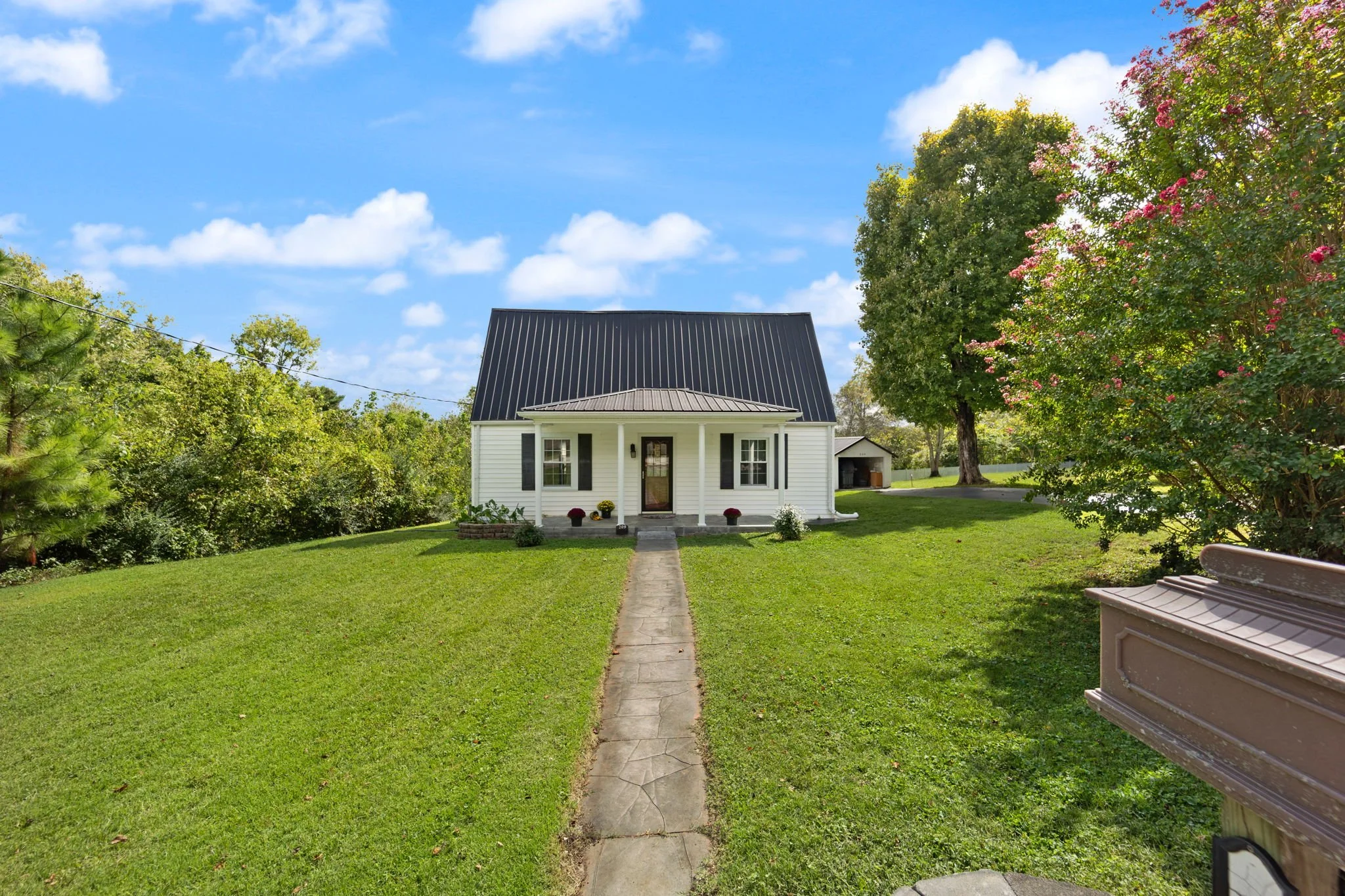 A small white house with black shutters and a black metal roof, surrounded by a well-kept lawn, trees, and shrubbery, under a partly cloudy blue sky.
