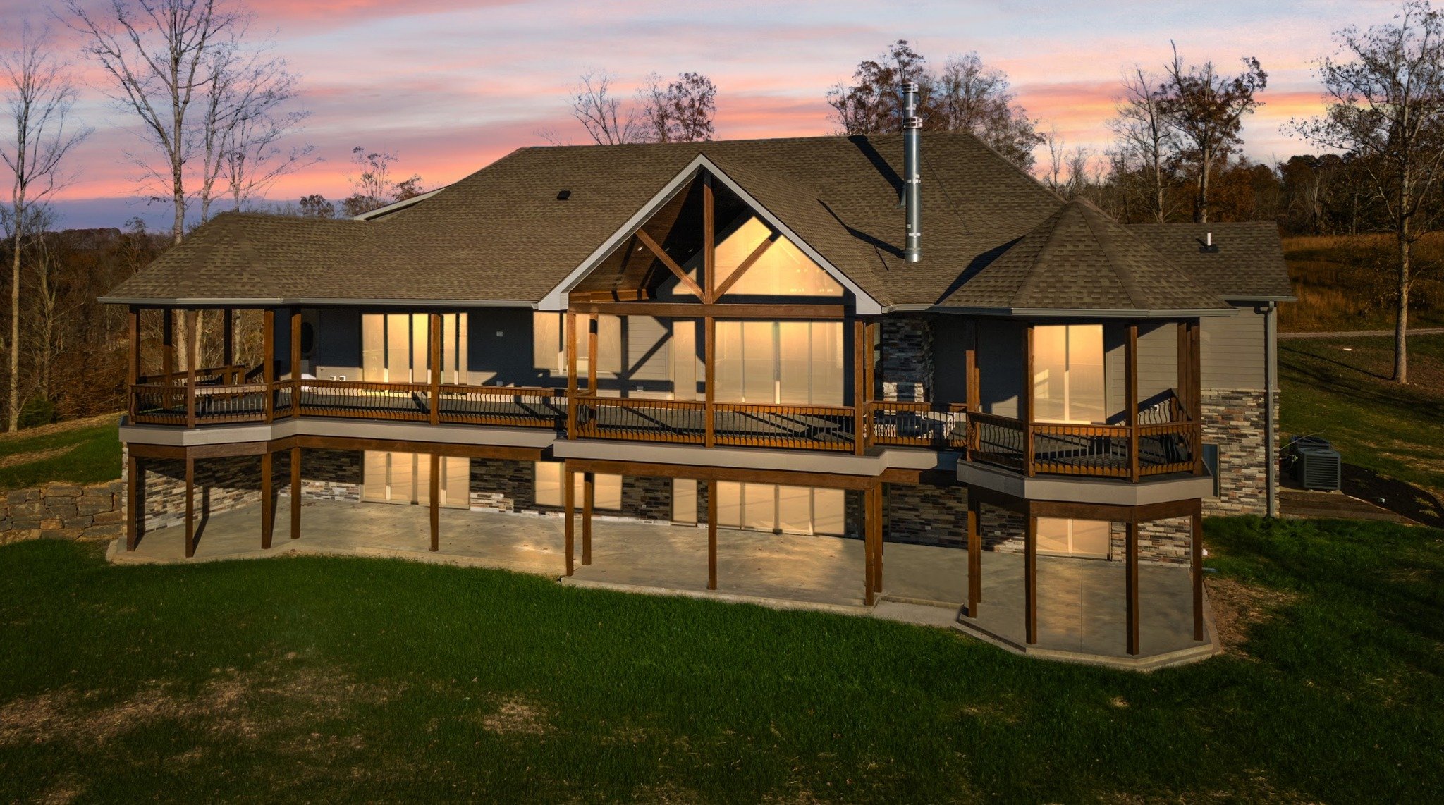 A modern house with large windows and a spacious wooden deck overlooking a grassy area during sunset.