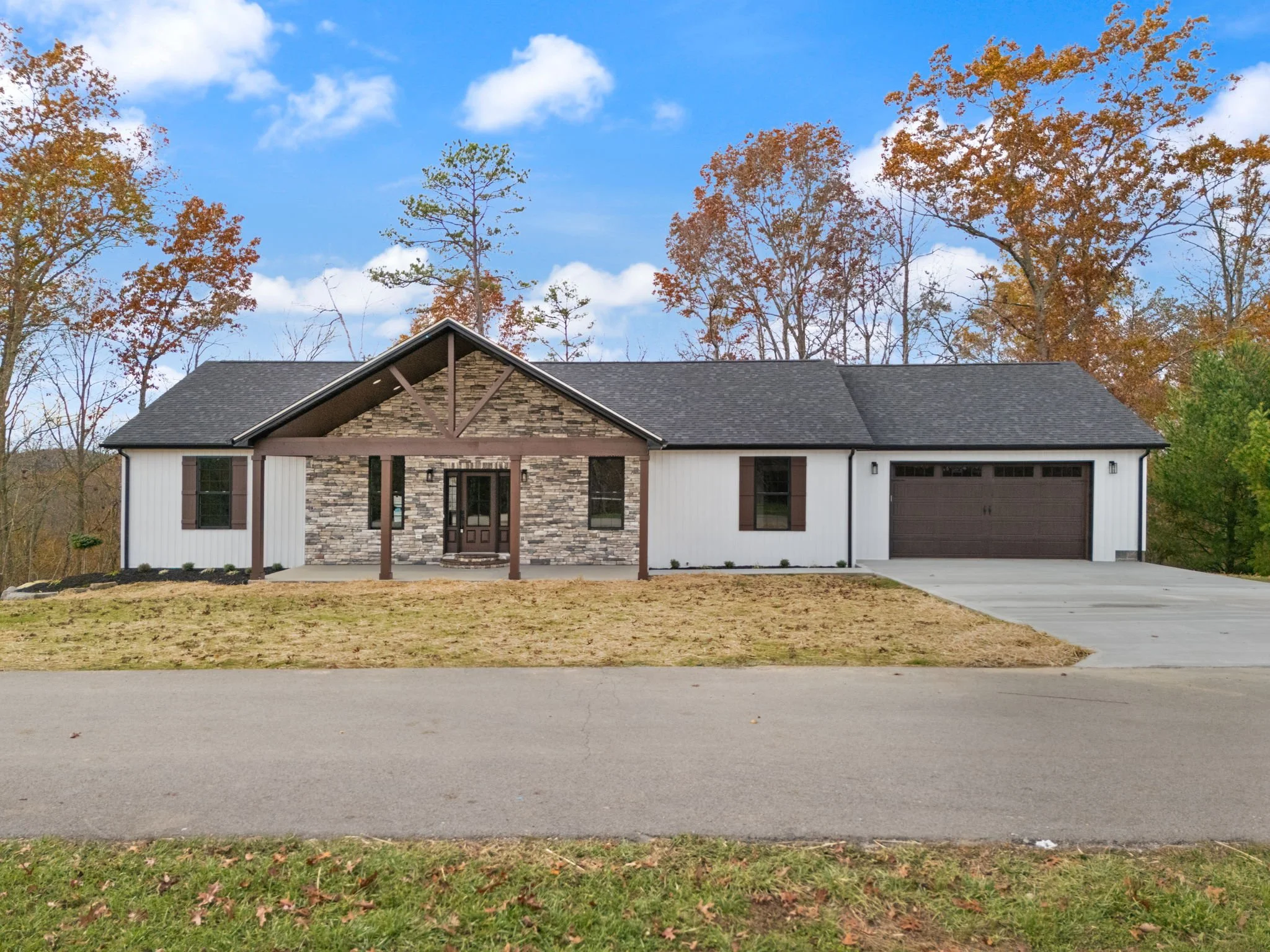 Single-story house with a combination of stone and white siding exterior, dark brown front door, attached garage, and a front slightly sloped lawn with a driveway, surrounded by trees with autumn foliage.