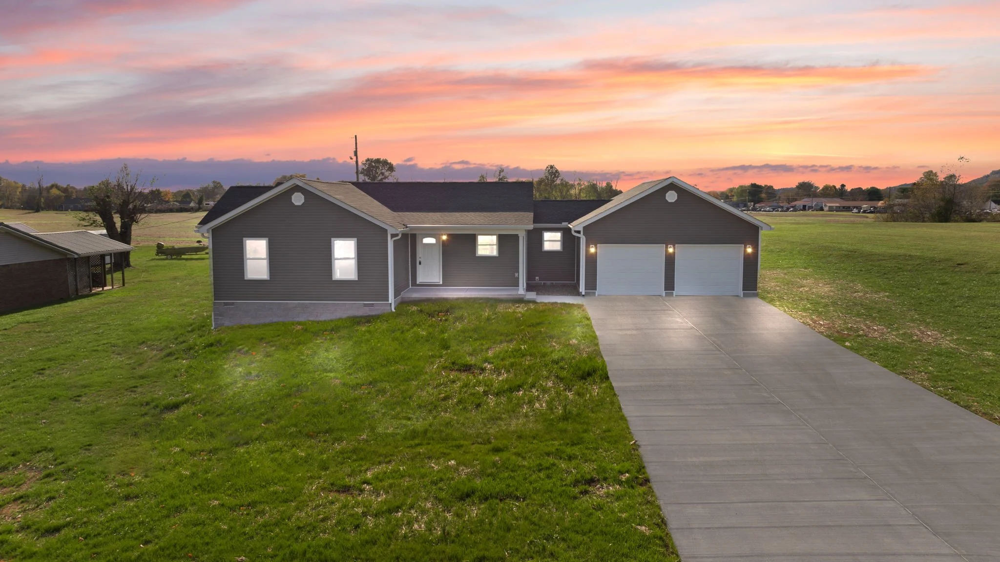 Modern single-family house with gray siding, three garage doors, and a driveway, set against a colorful sunset sky with green lawn and open fields.