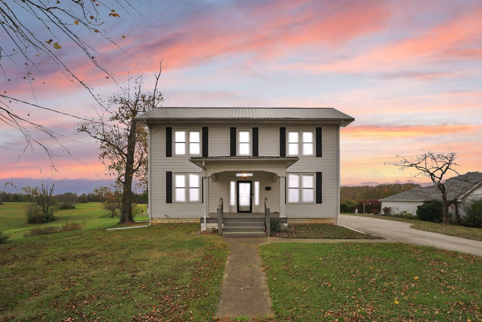 A two-story house with white siding, black shutters, a covered front porch, and a concrete walkway leading to the front steps, set against a colorful sunset sky with pink, orange, and purple clouds, and surrounded by a grassy yard with a few trees.