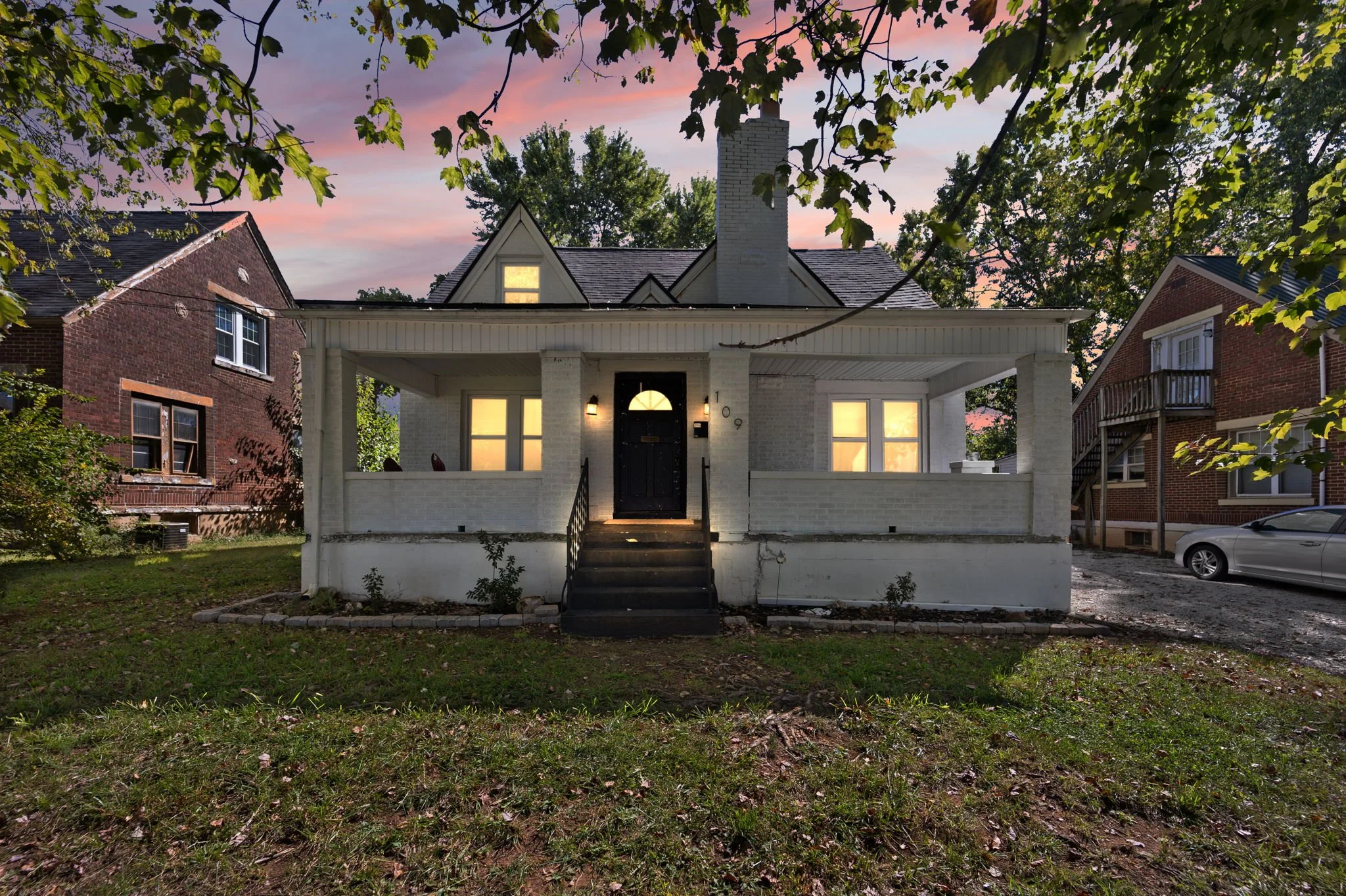 White painted house with front porch, black door, and lit windows, during sunset with pink and purple sky, neighboring brick houses, and green trees.