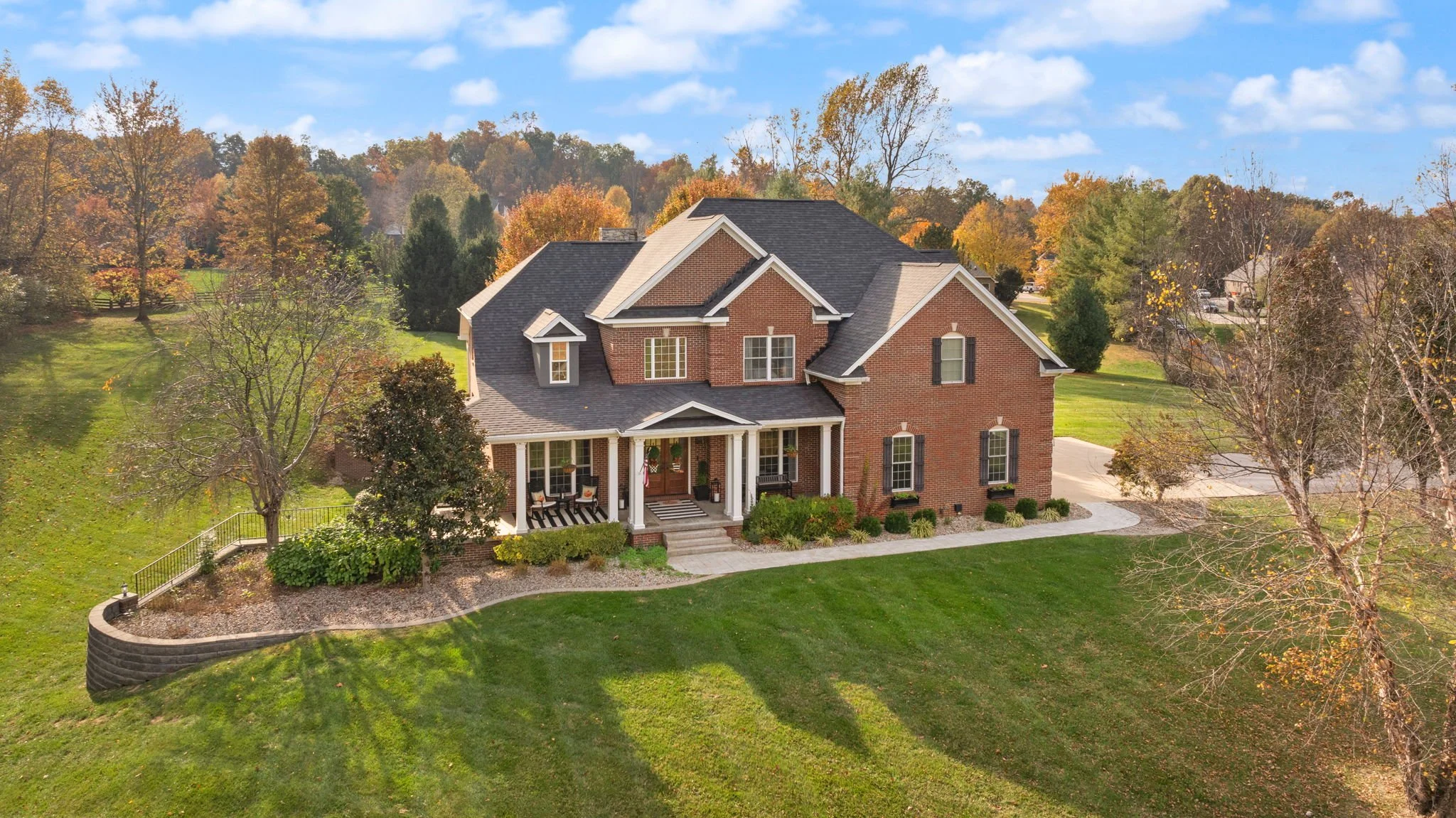 Large brick house with a front porch, surrounded by trees and green lawn, during fall with colorful foliage and clear blue sky.