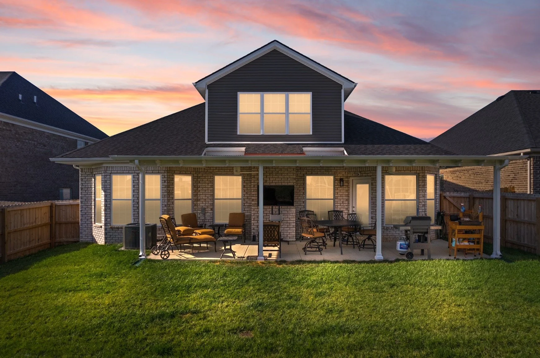 Backyard patio at sunset with outdoor furniture, grill, and a TV mounted on house wall, enclosed by a wooden fence.