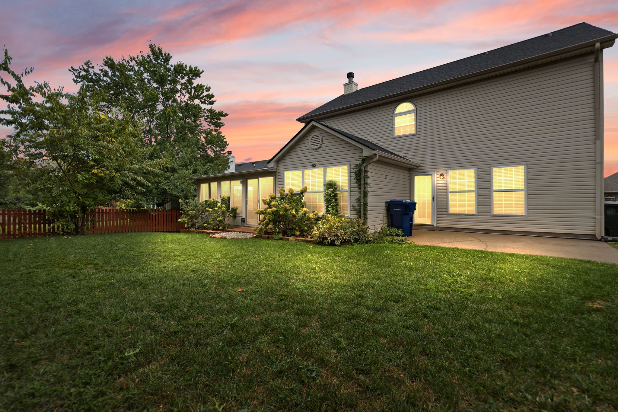 View of a suburban house at sunset with lit windows, a backyard with green grass, trees, and a fence.