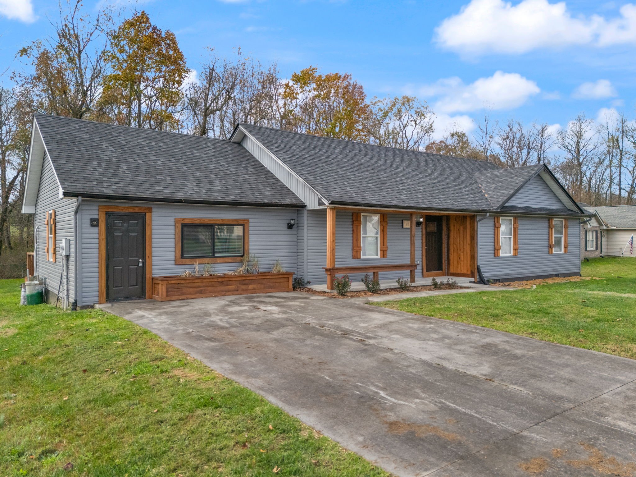 Single-story house with gray siding, black front door, and wooden accents, surrounded by a lawn and trees with fall foliage.