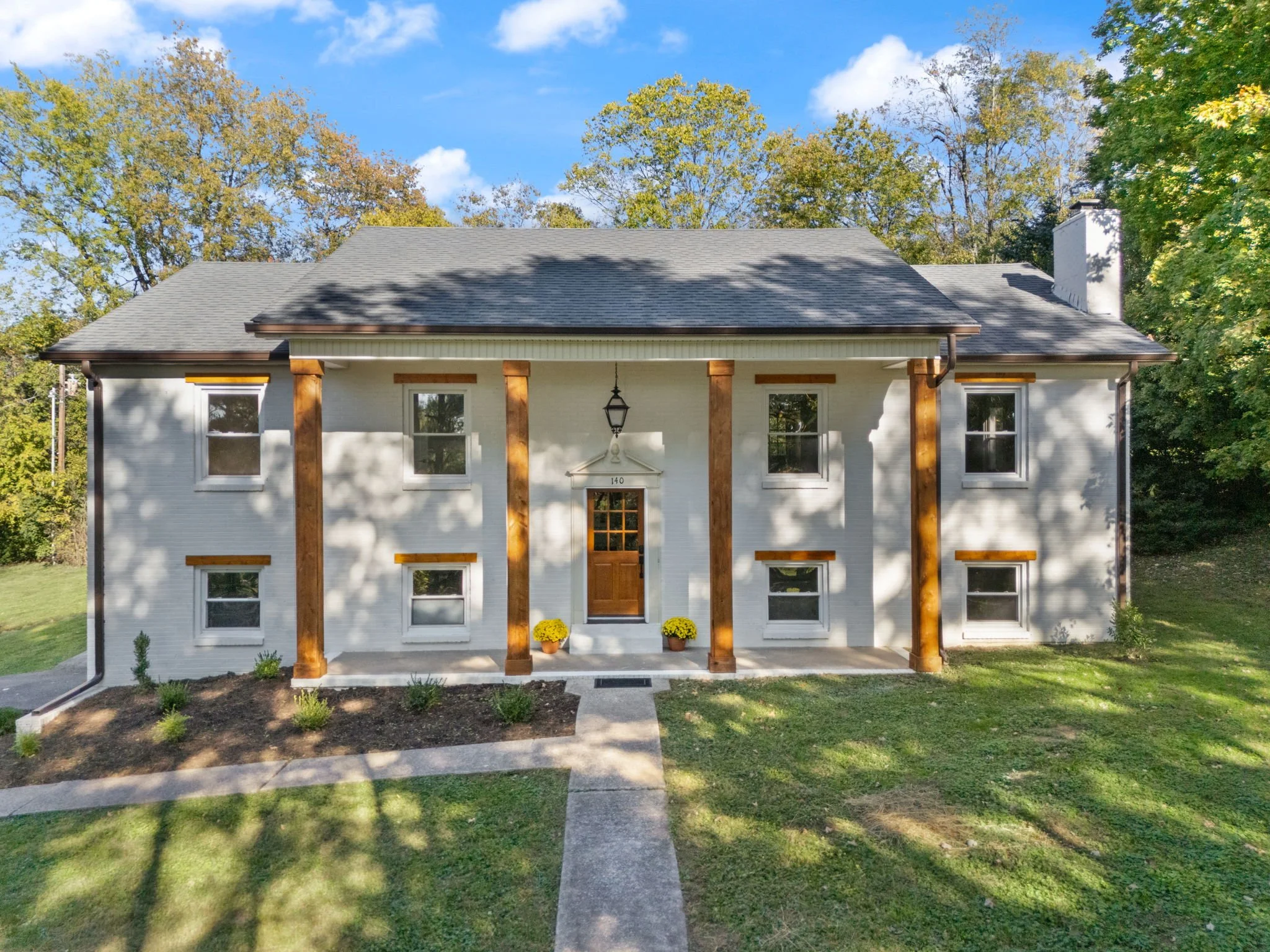White two-story house with wooden columns, front porch, and a dark gray roof, surrounded by green trees and lawn, under partly cloudy sky.