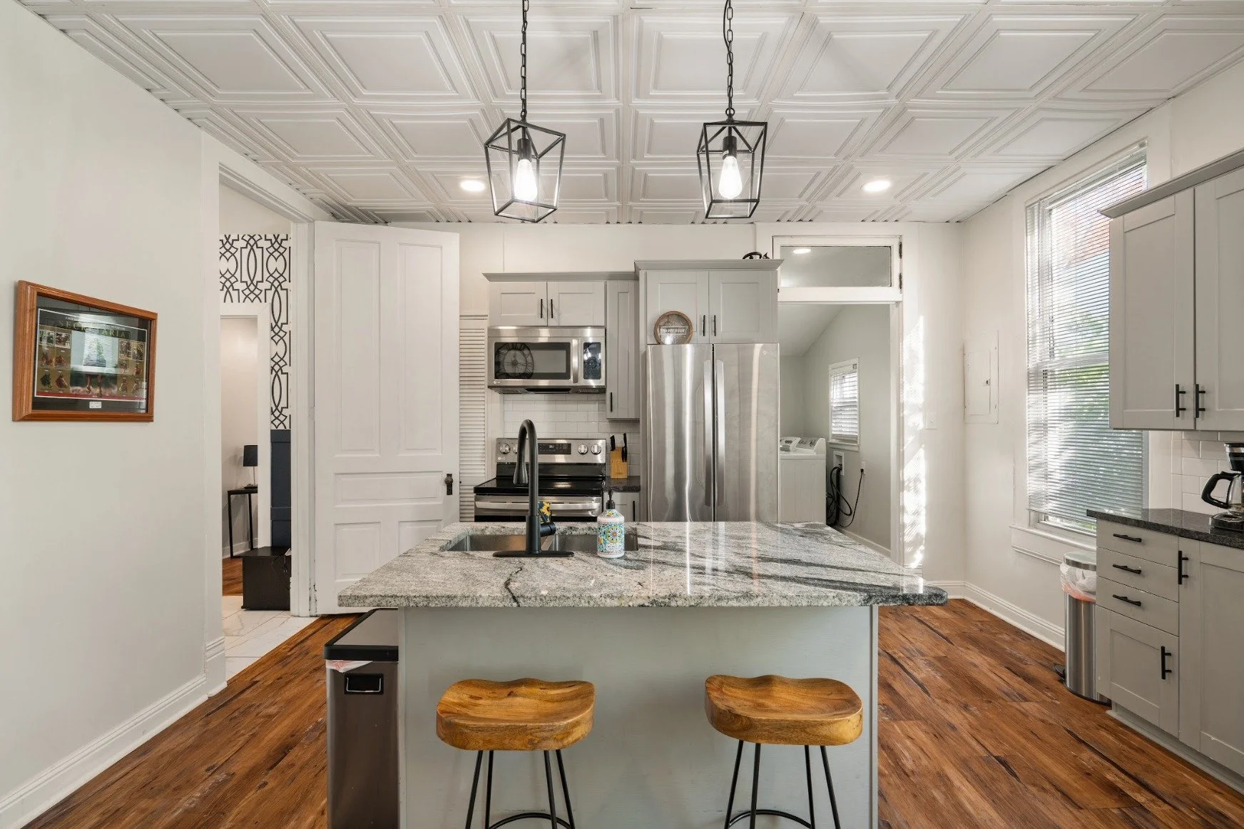 Modern kitchen with white cabinets, stainless steel appliances, granite island with two wooden stools, wood flooring, and hanging pendant lights.