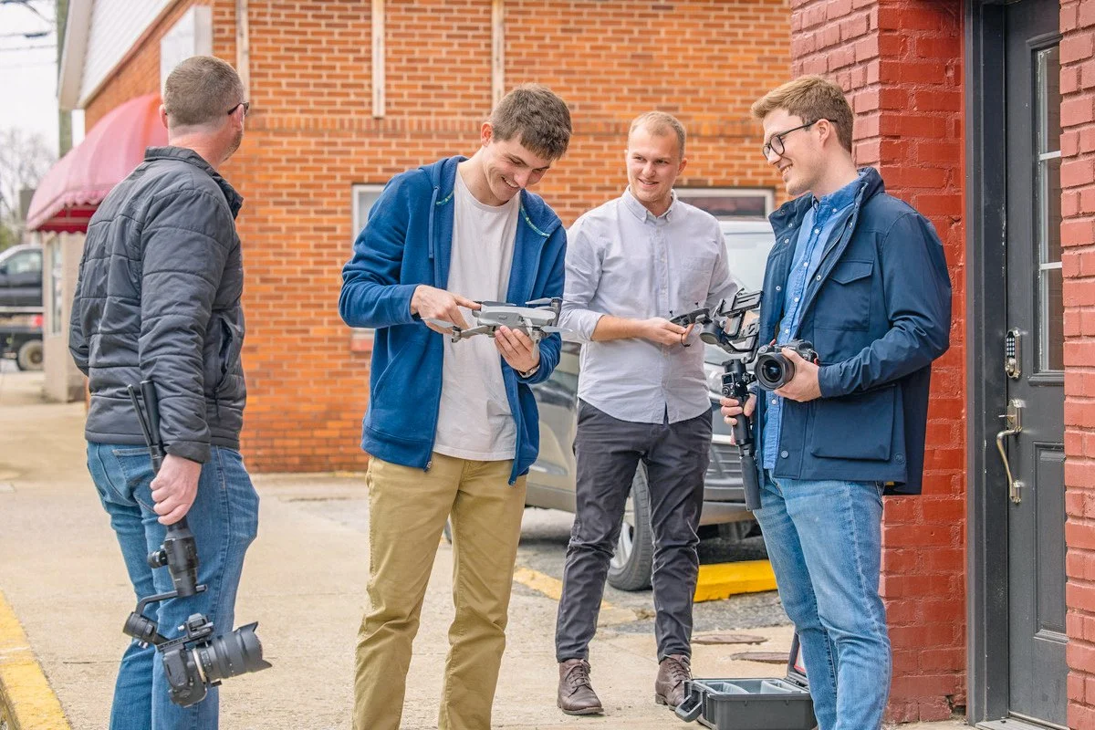 Four men standing outside a brick building, engaged in a conversation about their photography gear. One man holds a camera, another is holding a drone, and the others have camera equipment.