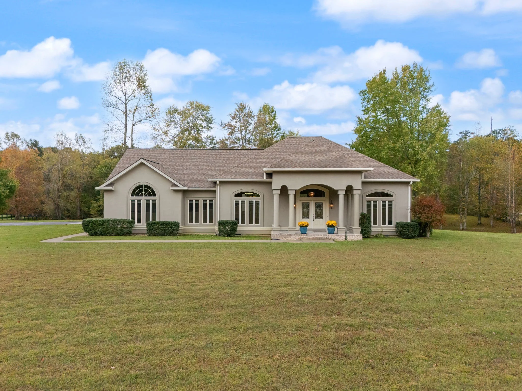 Large, beige stucco house with arched windows, a front porch with two columns, and potted yellow flowers, surrounded by green lawn and trees with autumn foliage under a partly cloudy sky.