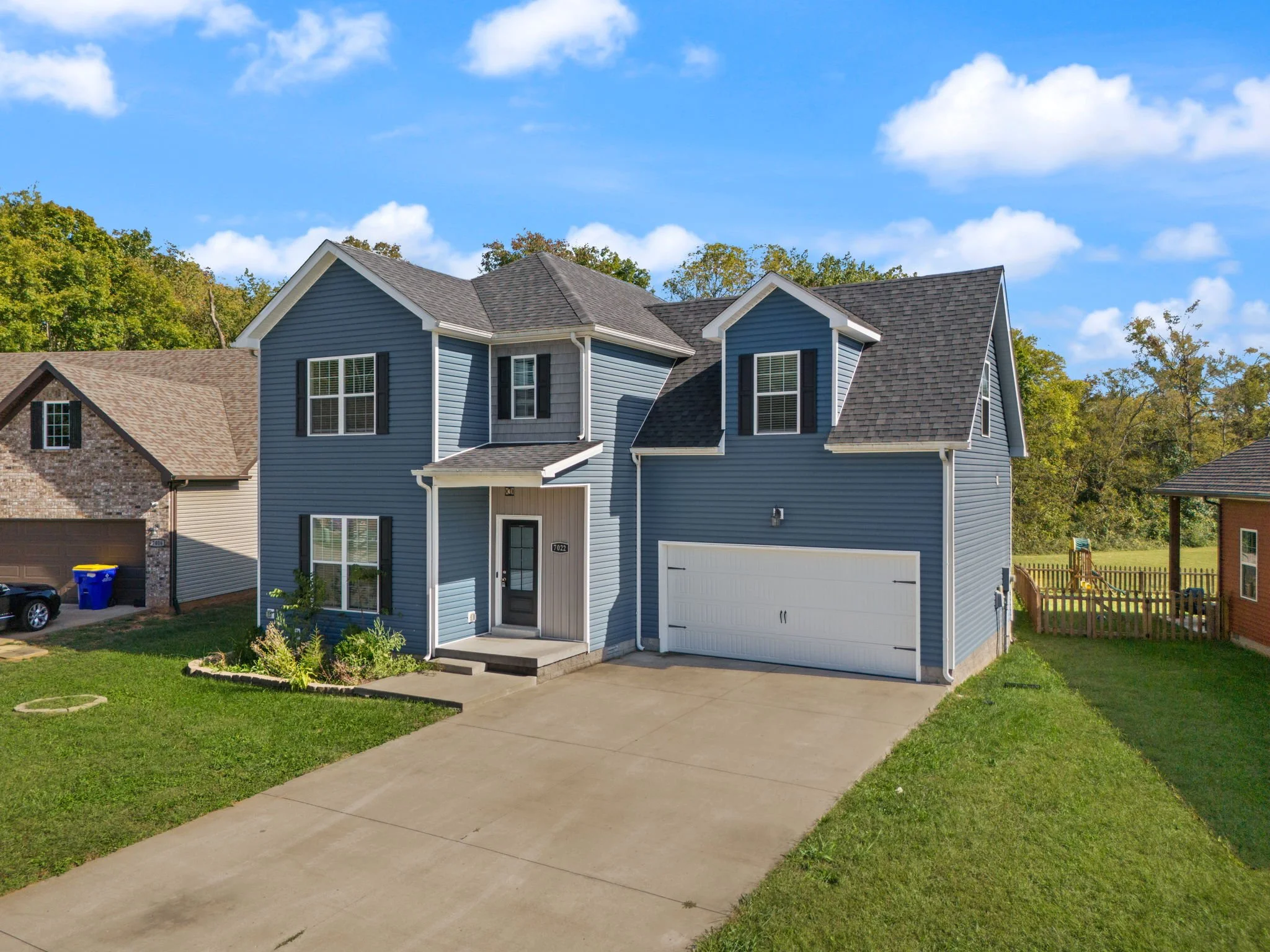 Front view of a blue two-story house with a white garage door and a small concrete porch, surrounded by green lawns and neighboring houses, under a partly cloudy sky.