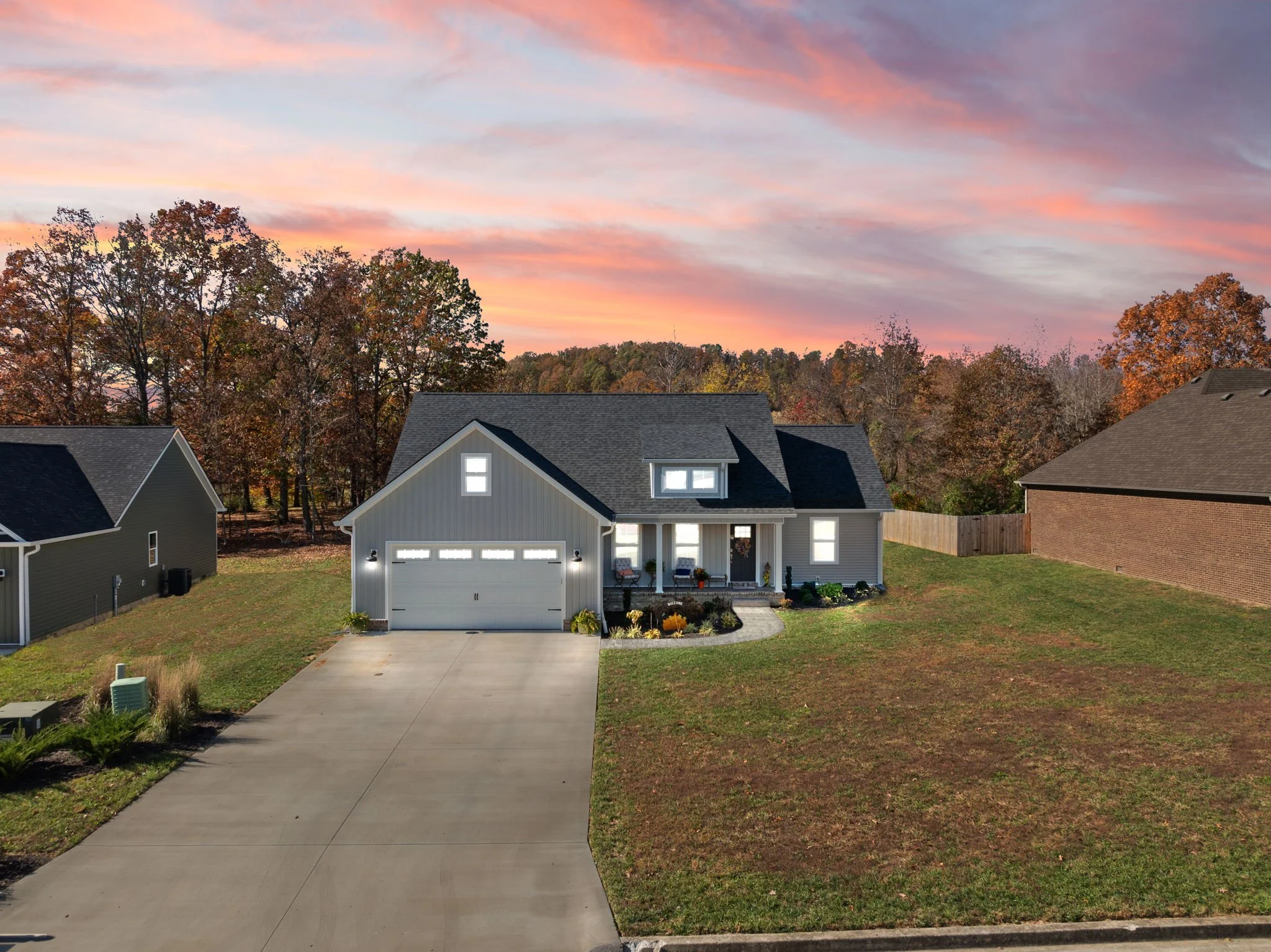 A modern two-story house with gray siding, white garage door, and front porch with chairs, set against a backdrop of autumn trees and a colorful sunset sky.