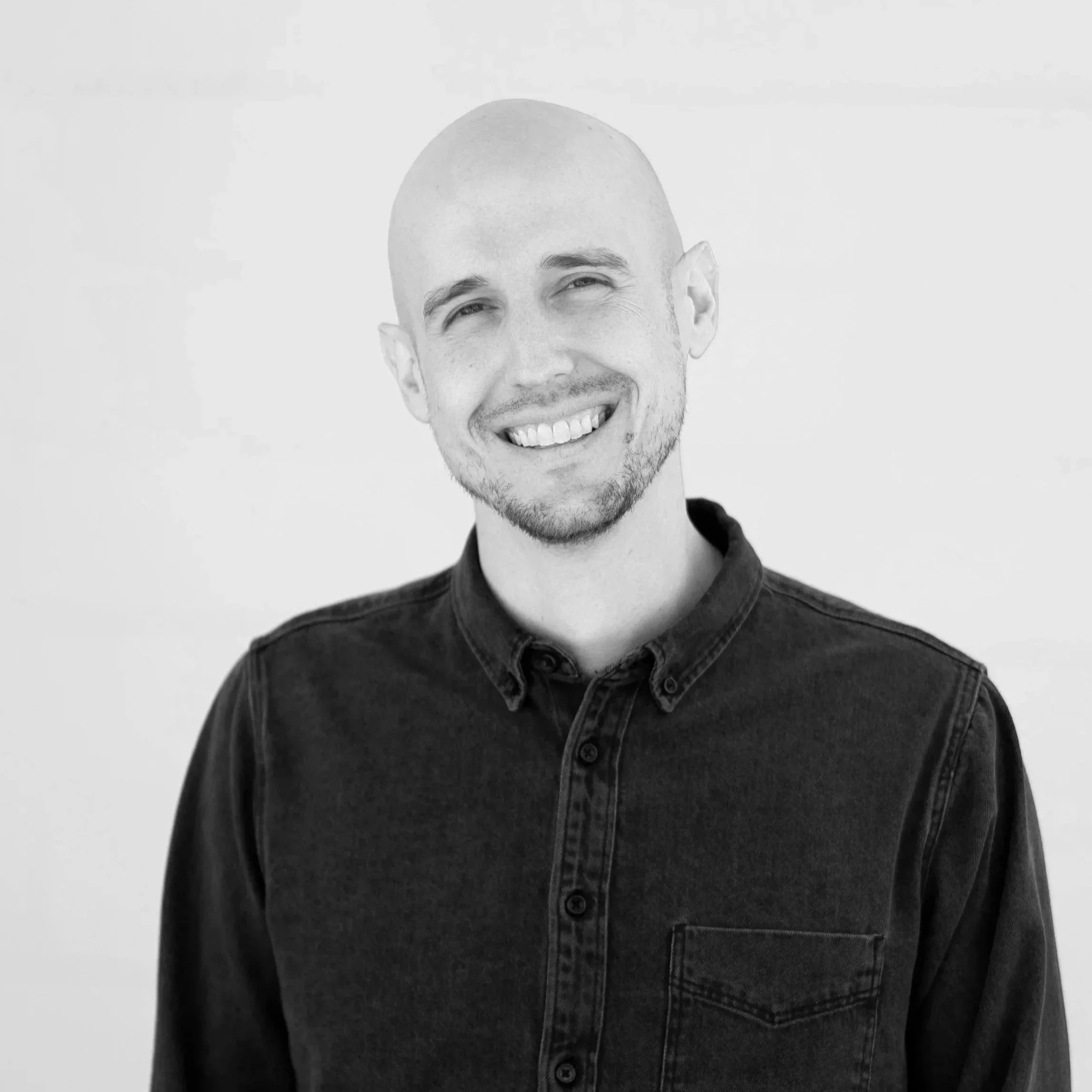 A smiling, bald man with a goatee, wearing a black button-up shirt, standing against a plain white background.