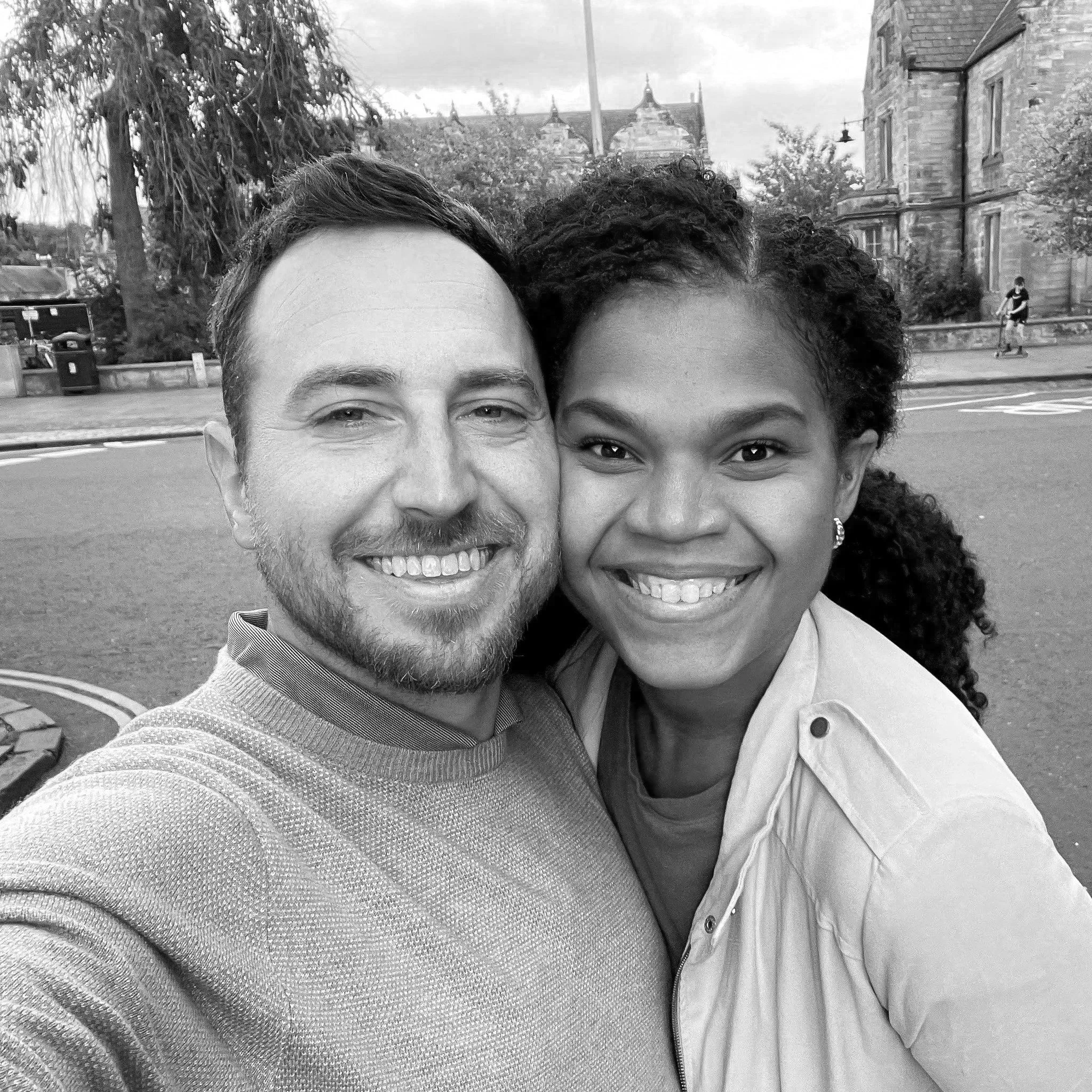 A black and white photograph of a smiling couple, a man and a woman, close together outdoors with a blurred natural background.